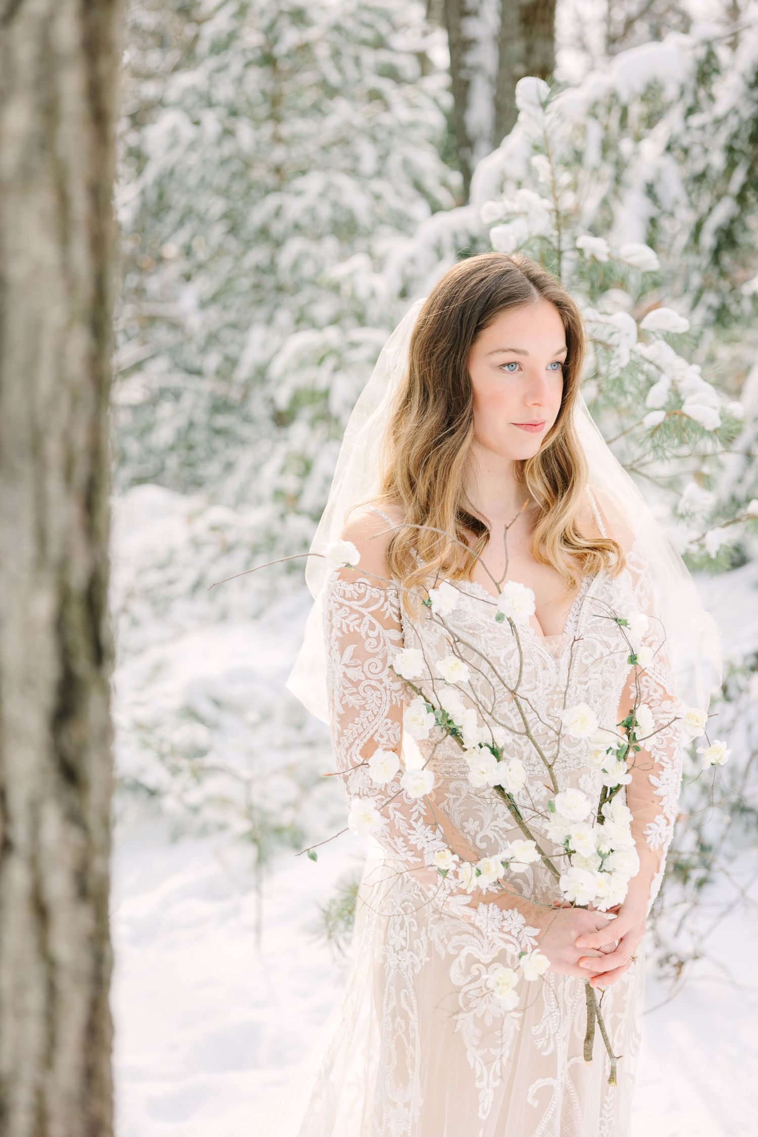 Elegant bride wearing lace wedding dress and veil holding white flowers among snow-covered pine trees.