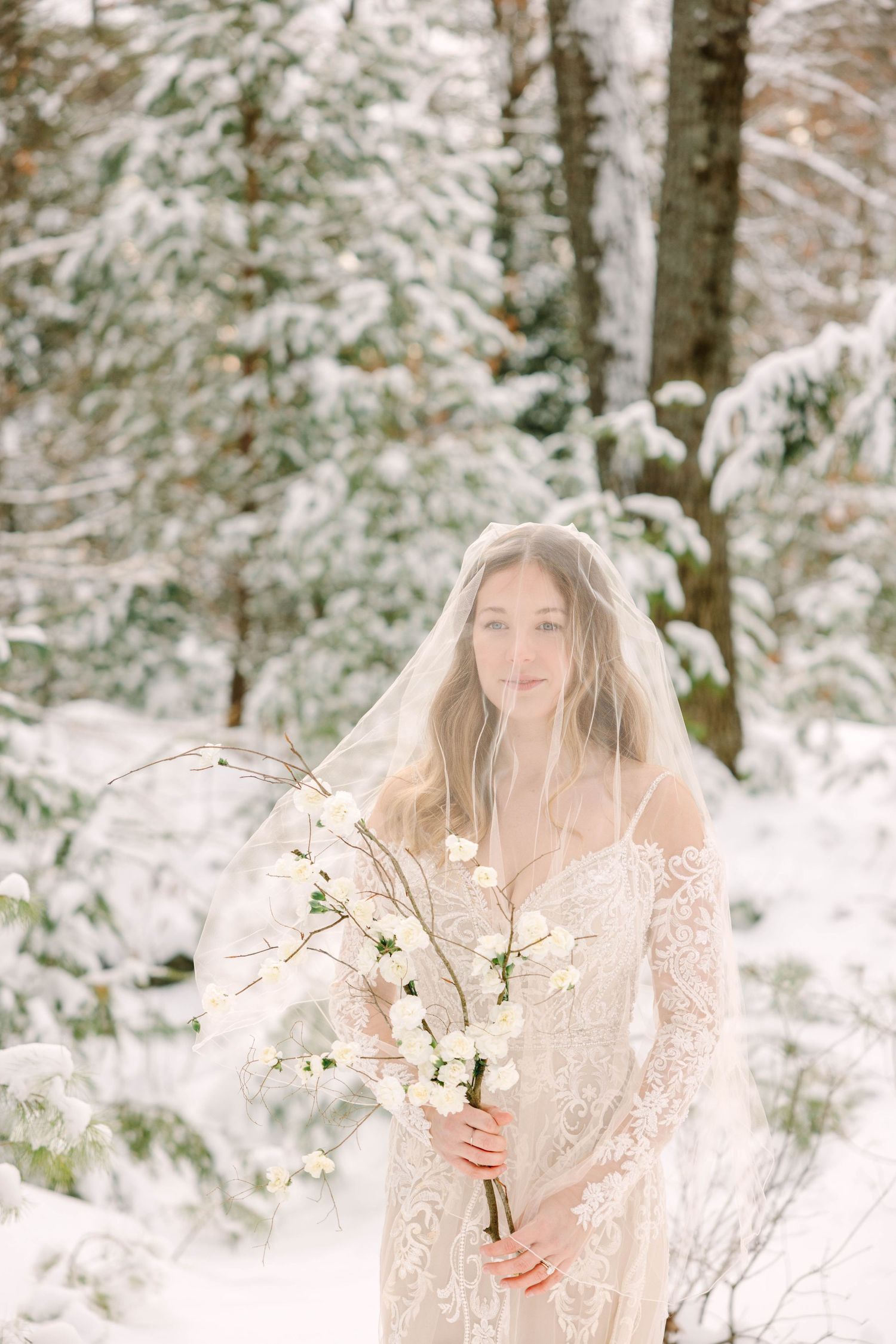 Bride wearing lace gown and flowing veil holds white floral bouquet among snow-dusted pine trees.