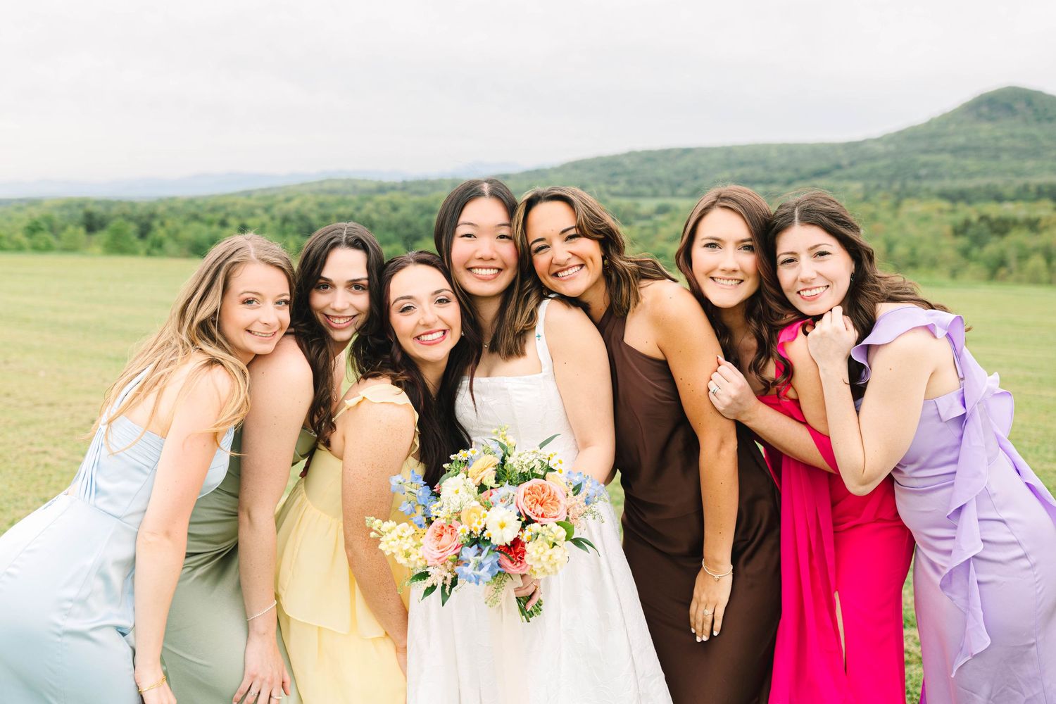 Bride in white gown holds colorful bouquet surrounded by six bridesmaids in pastel and bold dresses outdoors.