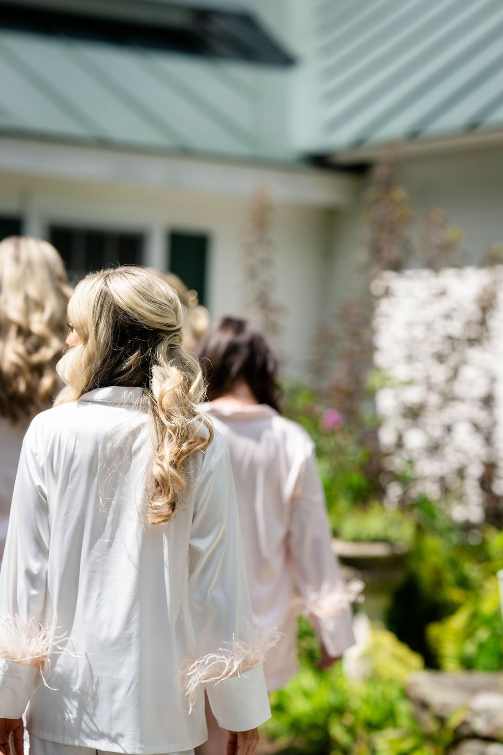 Bridesmaids in white feather-trimmed robes walking outdoors near a garden with blooming flowers on a sunny day.