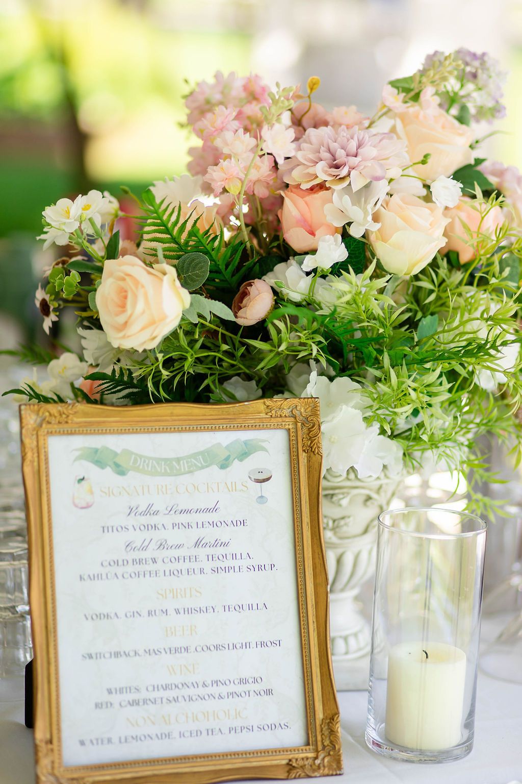 Elegant wedding table with gold-framed drink menu, pink roses, and white candle centerpiece.