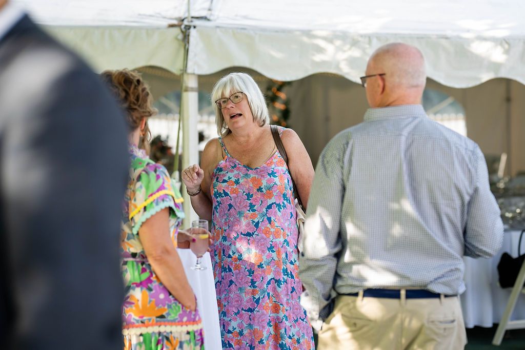 Woman in floral dress laughs joyfully with guests under white tent at outdoor wedding.