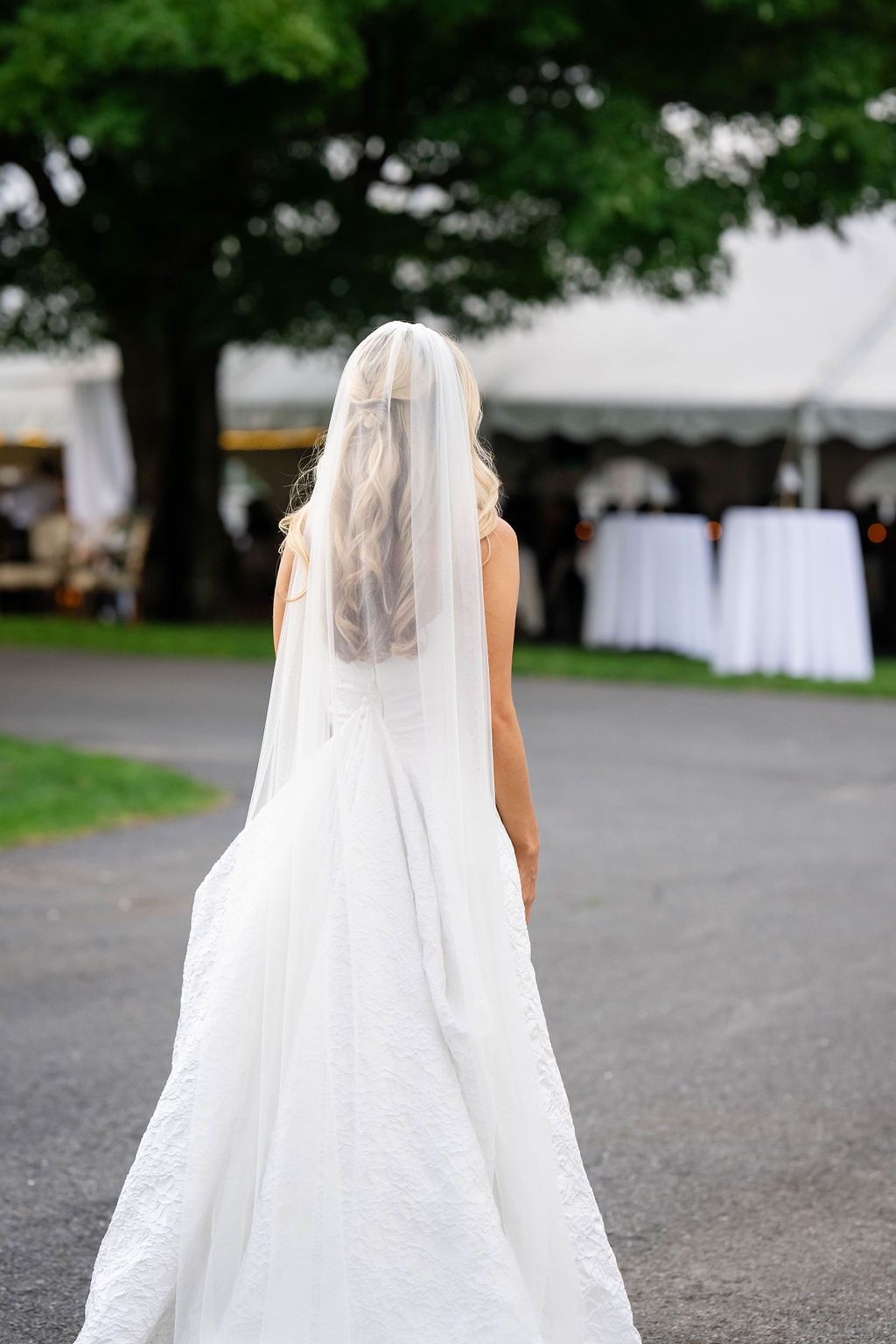 Bride in white lace wedding gown and long veil walking toward reception tent from behind.