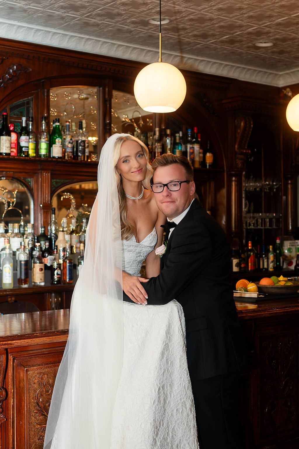 Bride and groom pose together at vintage wooden bar with bottle display at wedding venue.