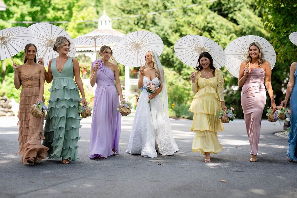Bride and bridesmaids in colorful pastel gowns holding white parasols walking outdoors at a summer wedding.