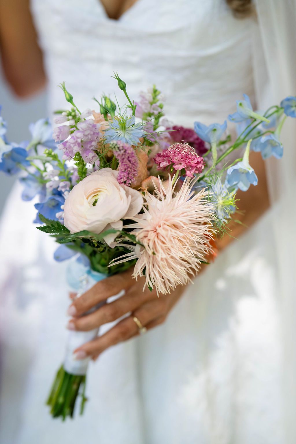 Bride holds a romantic pastel bouquet featuring ranunculus, blue delphinium, and pink blooms on wedding day.