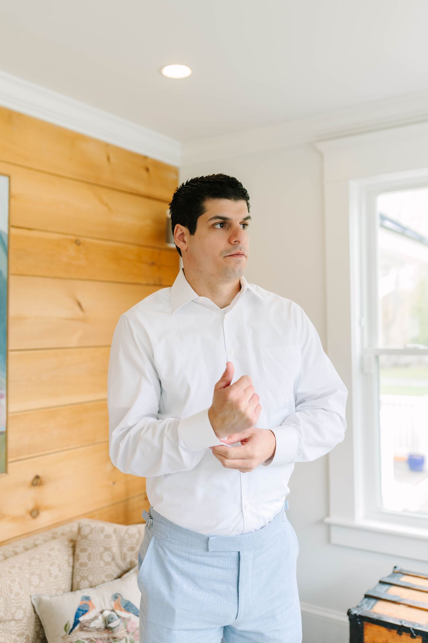 Groom in white dress shirt and light blue pants adjusting cufflinks indoors before wedding ceremony.