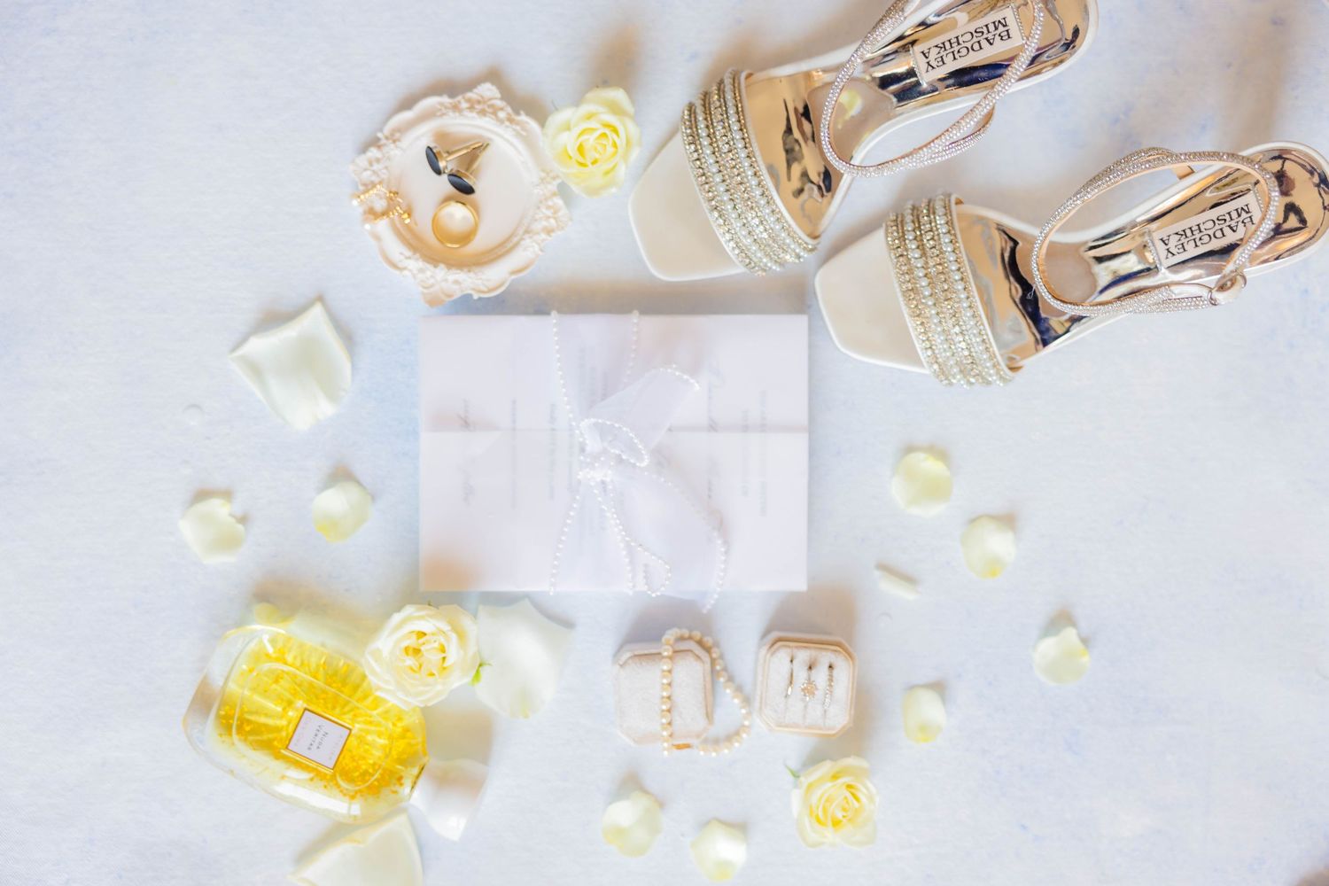 Elegant wedding flat lay with gold heels, white gift box, yellow roses, and perfume on a white background.