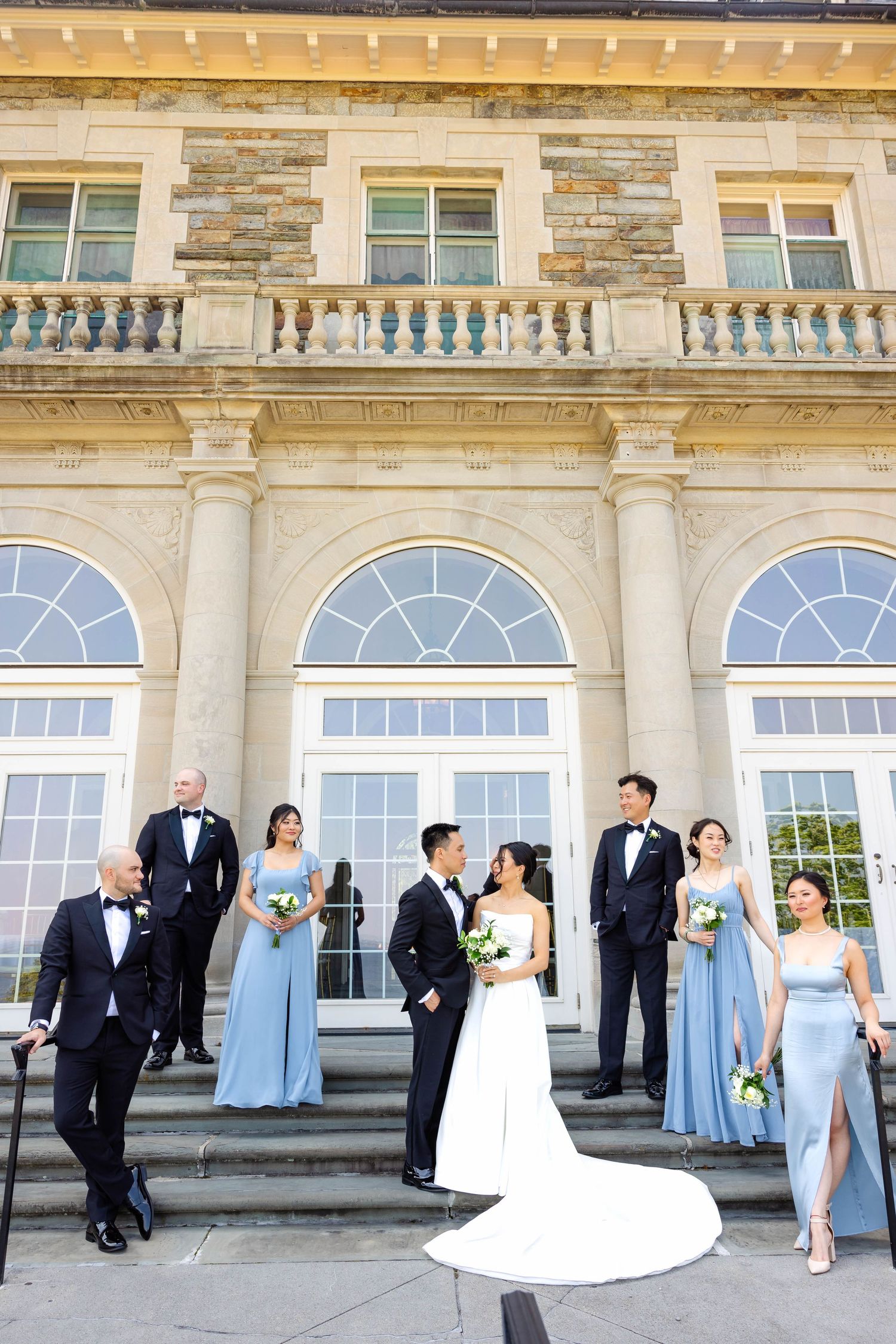 Wedding party in dusty blue gowns and black tuxedos poses on steps of a grand stone mansion.