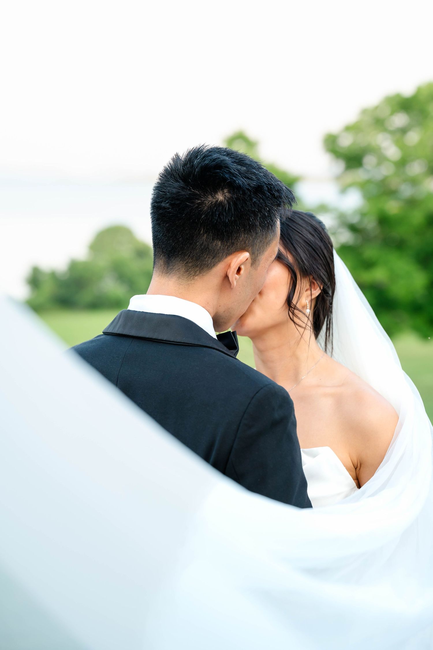 Romantic outdoor wedding kiss with bride in white strapless gown and flowing veil against lush green backdrop.