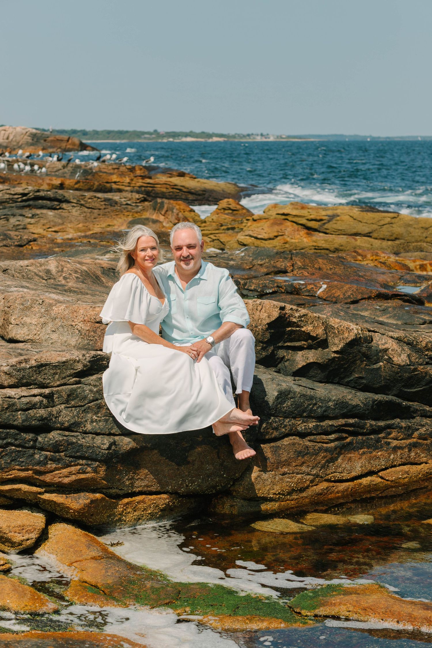 A smiling couple in white outfits sits on rocky coastal shore with ocean waves in the background on a sunny day.