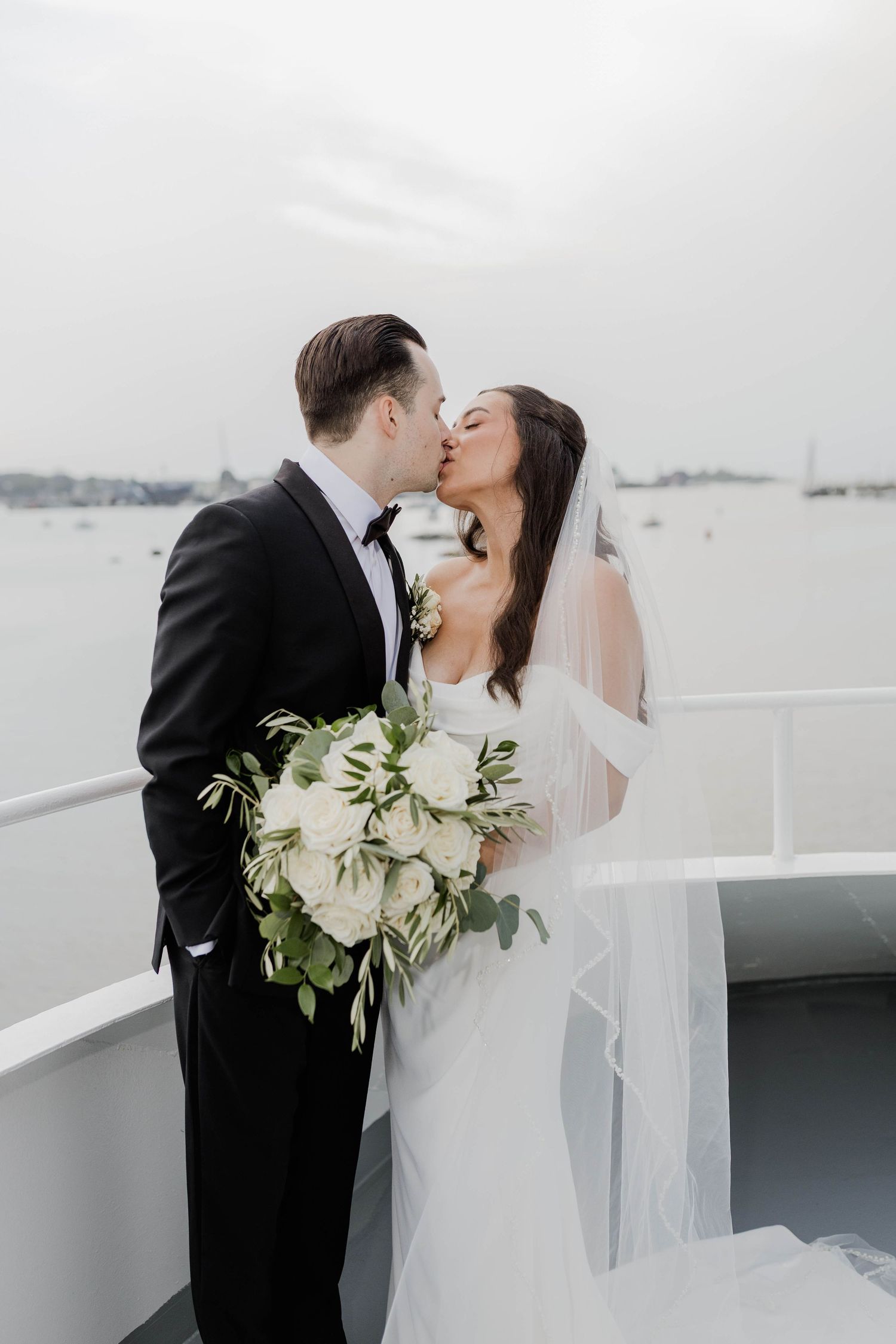 Bride and groom kissing on boat deck, bride holding white rose bouquet with greenery, veil flowing.