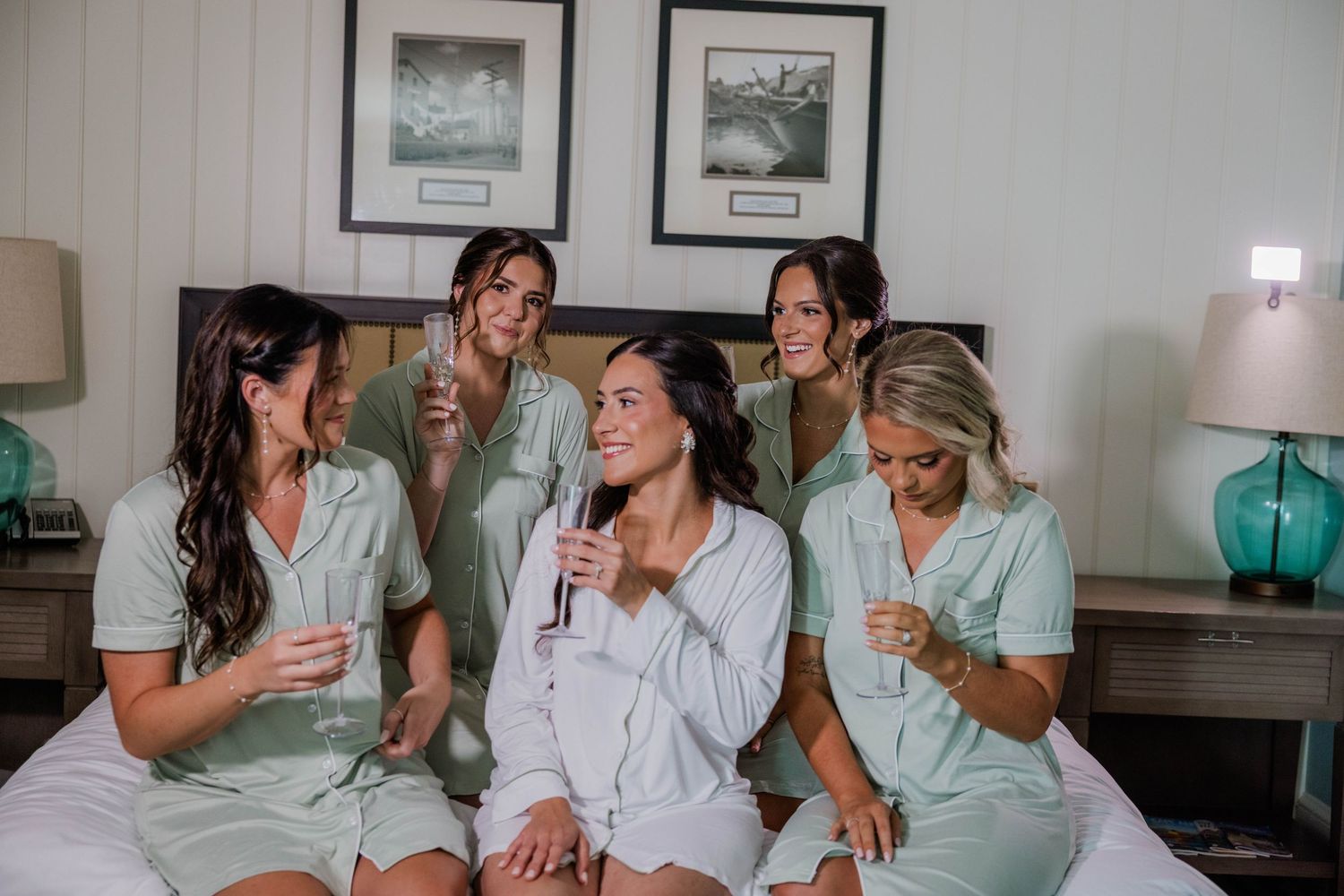 Bride and bridesmaids in matching sage green pajamas sitting on bed celebrating with champagne glasses.