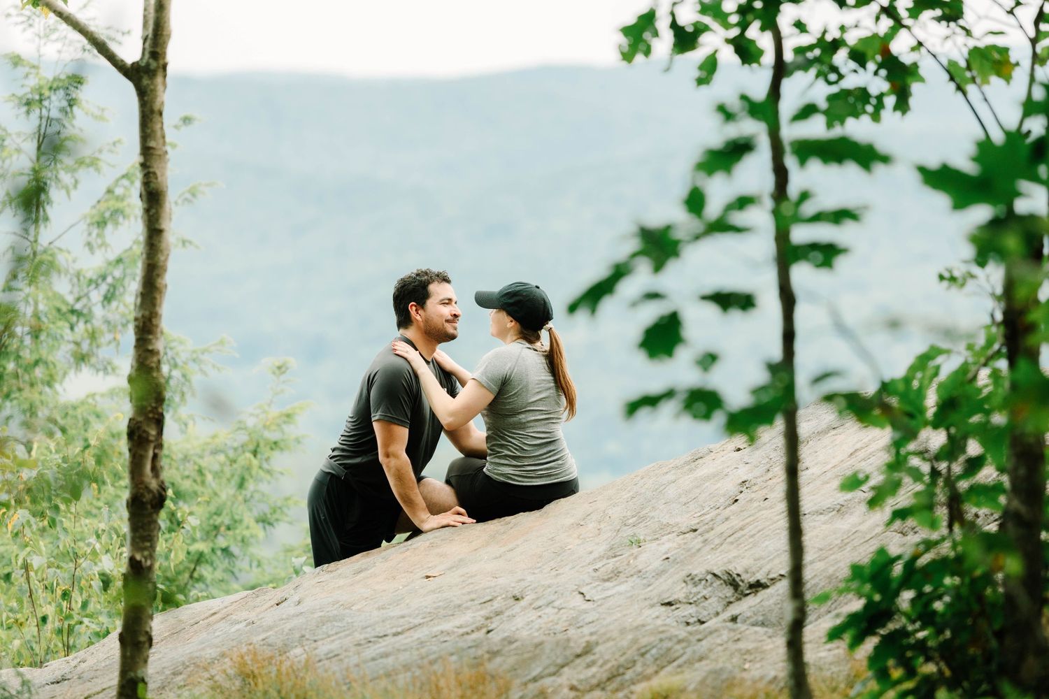 Couple sitting together on a rocky ledge enjoying scenic mountain views through green forest trees.