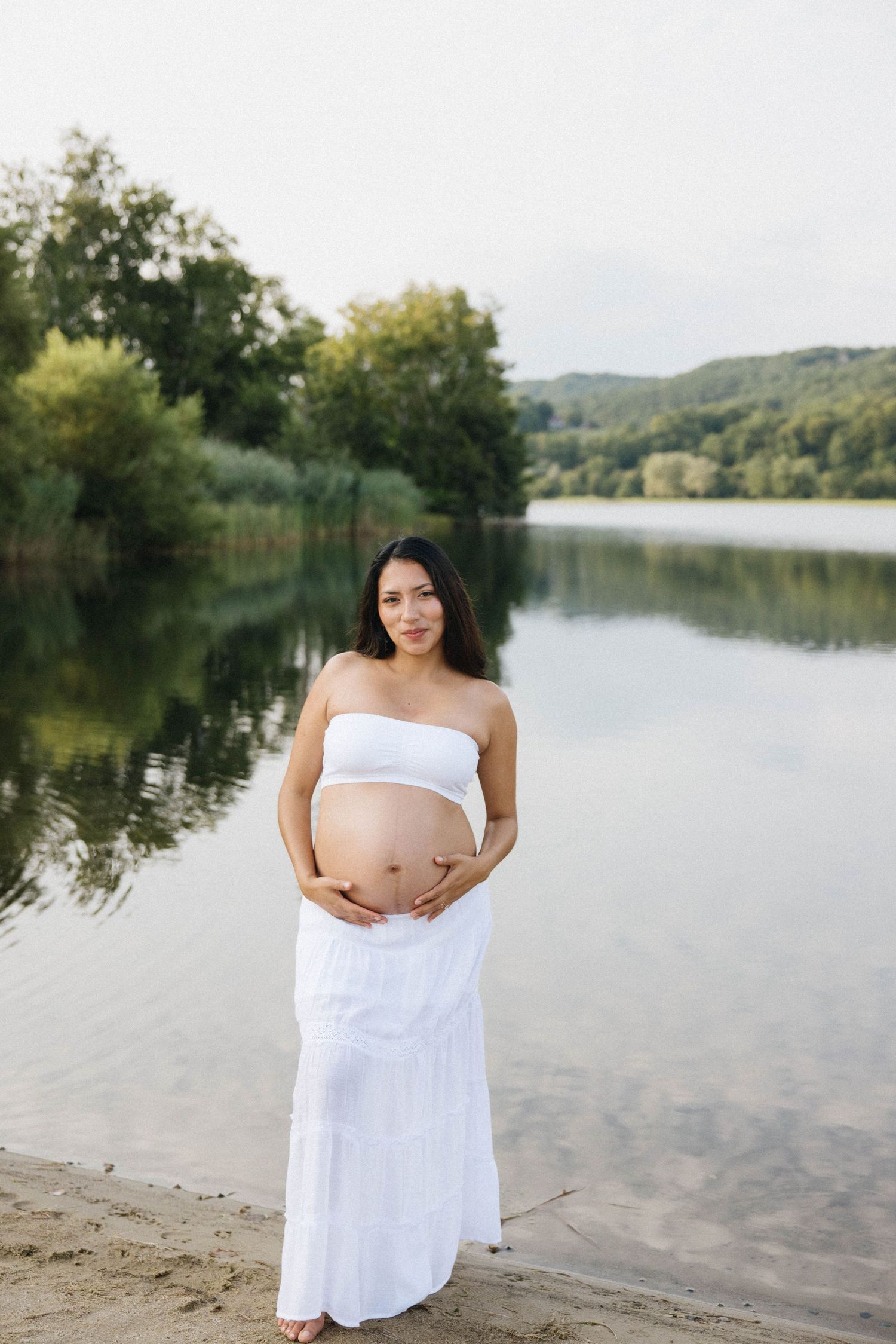Pregnant woman in white strapless top and skirt holding her baby bump by a serene lake surrounded by lush green trees.