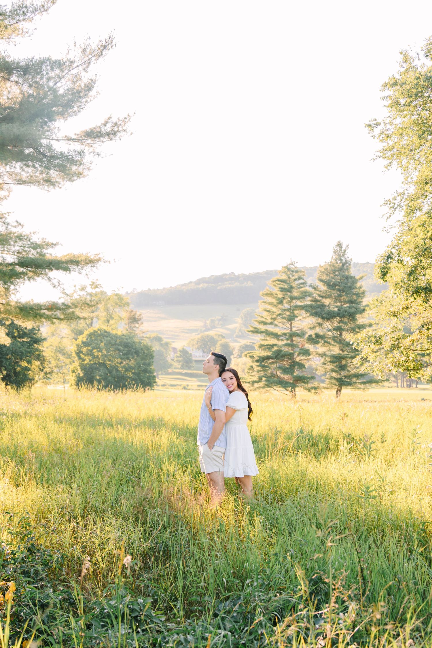 Couple standing together in a sunlit meadow surrounded by trees and rolling hills.