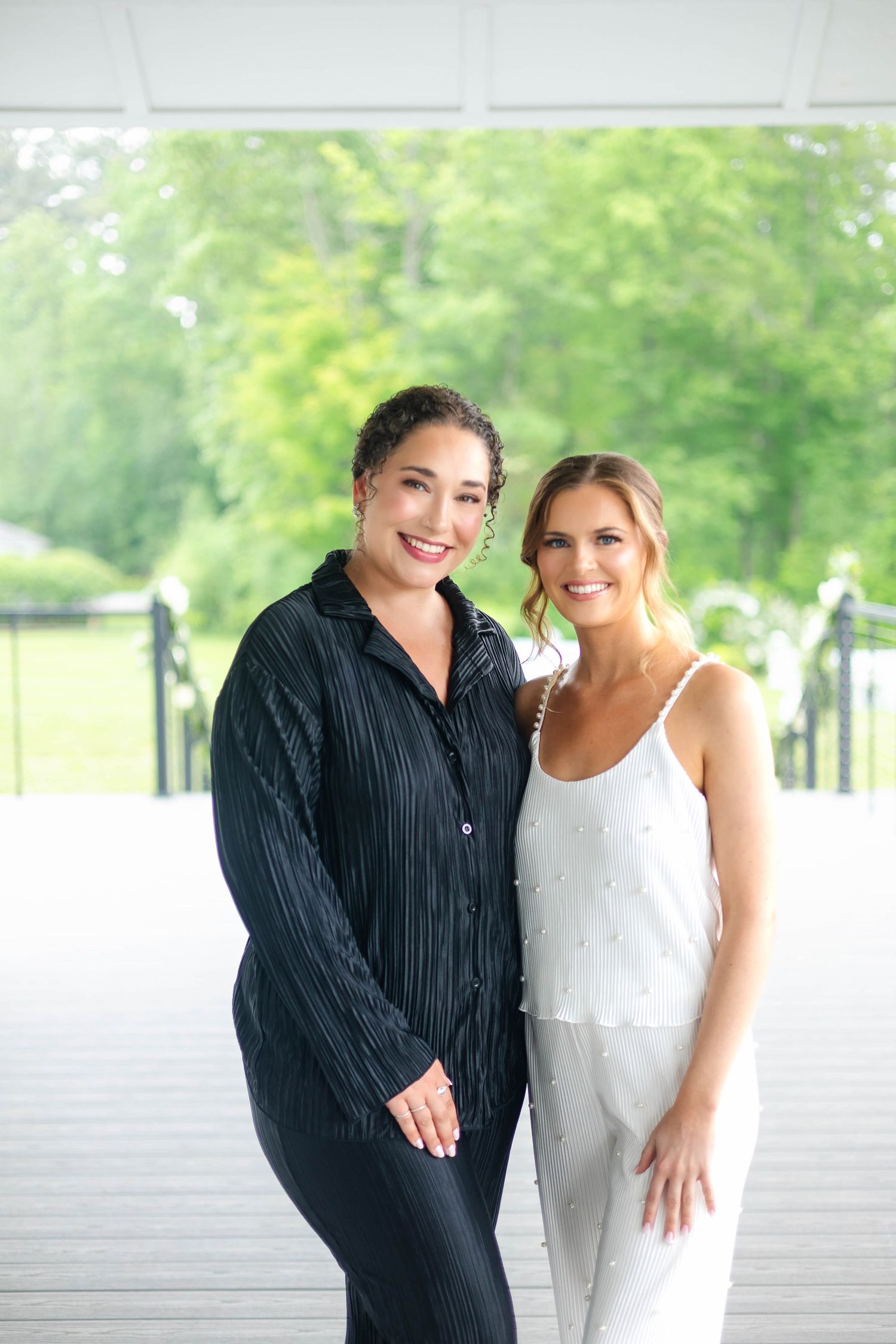 Bride in white camisole pajamas smiling with bridesmaid in black outfit on outdoor deck.