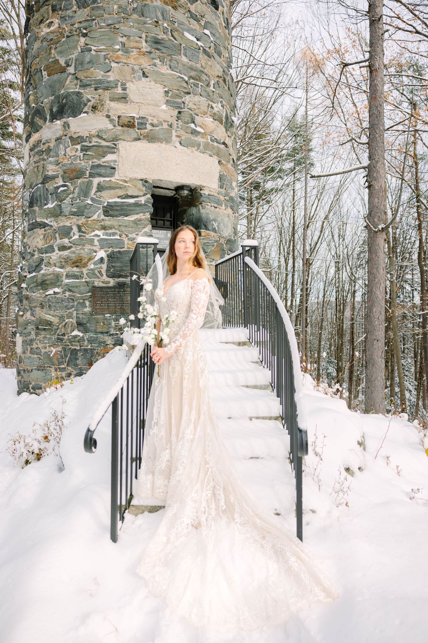Bride in long lace gown descends snowy stone tower steps holding white floral bouquet.