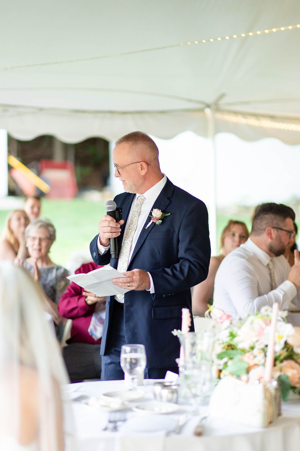 Father of bride delivering heartfelt speech at outdoor wedding reception under white tent.