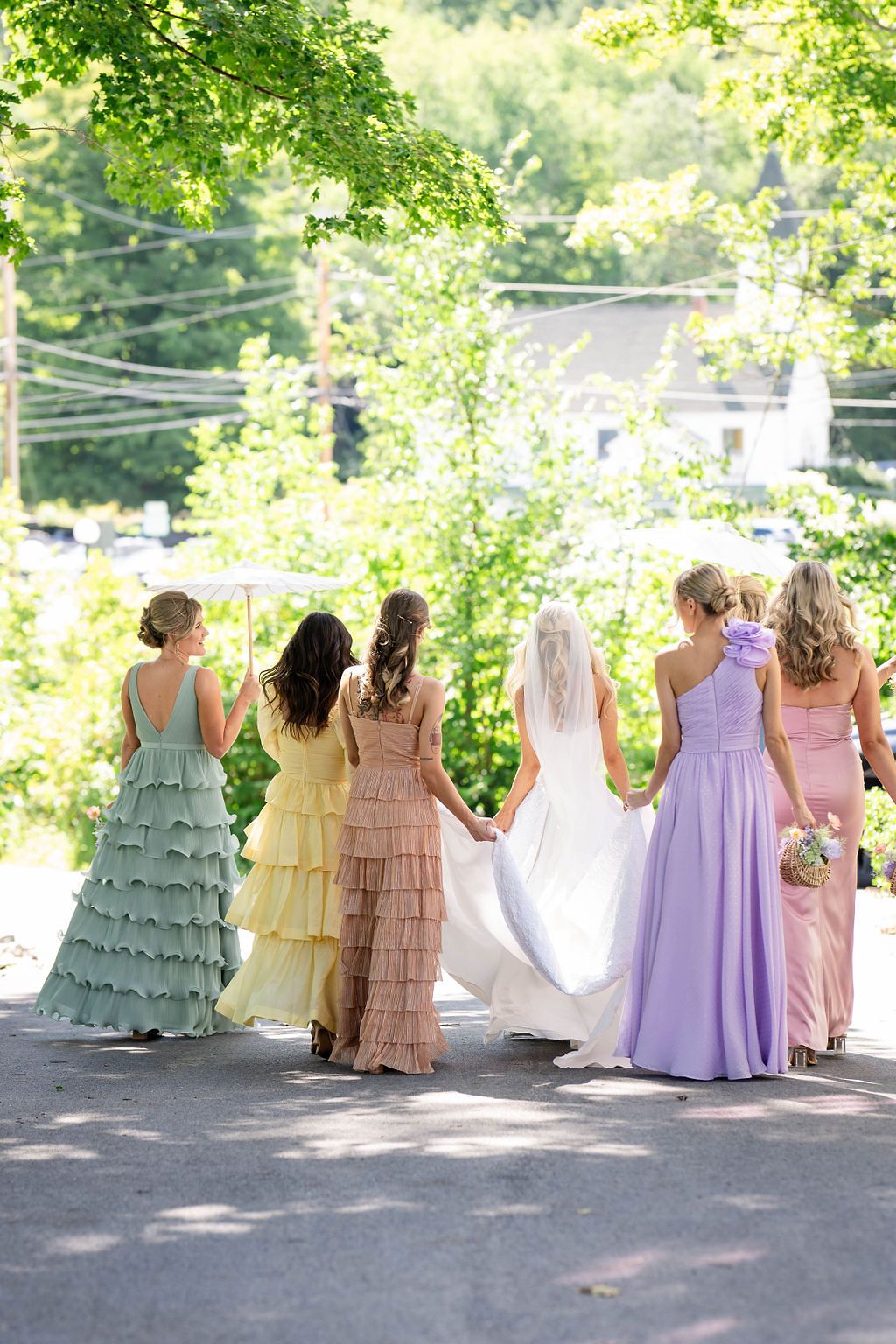 Back view of bride in white gown walking with bridesmaids in mint, yellow, peach, lavender, and pink dresses.
