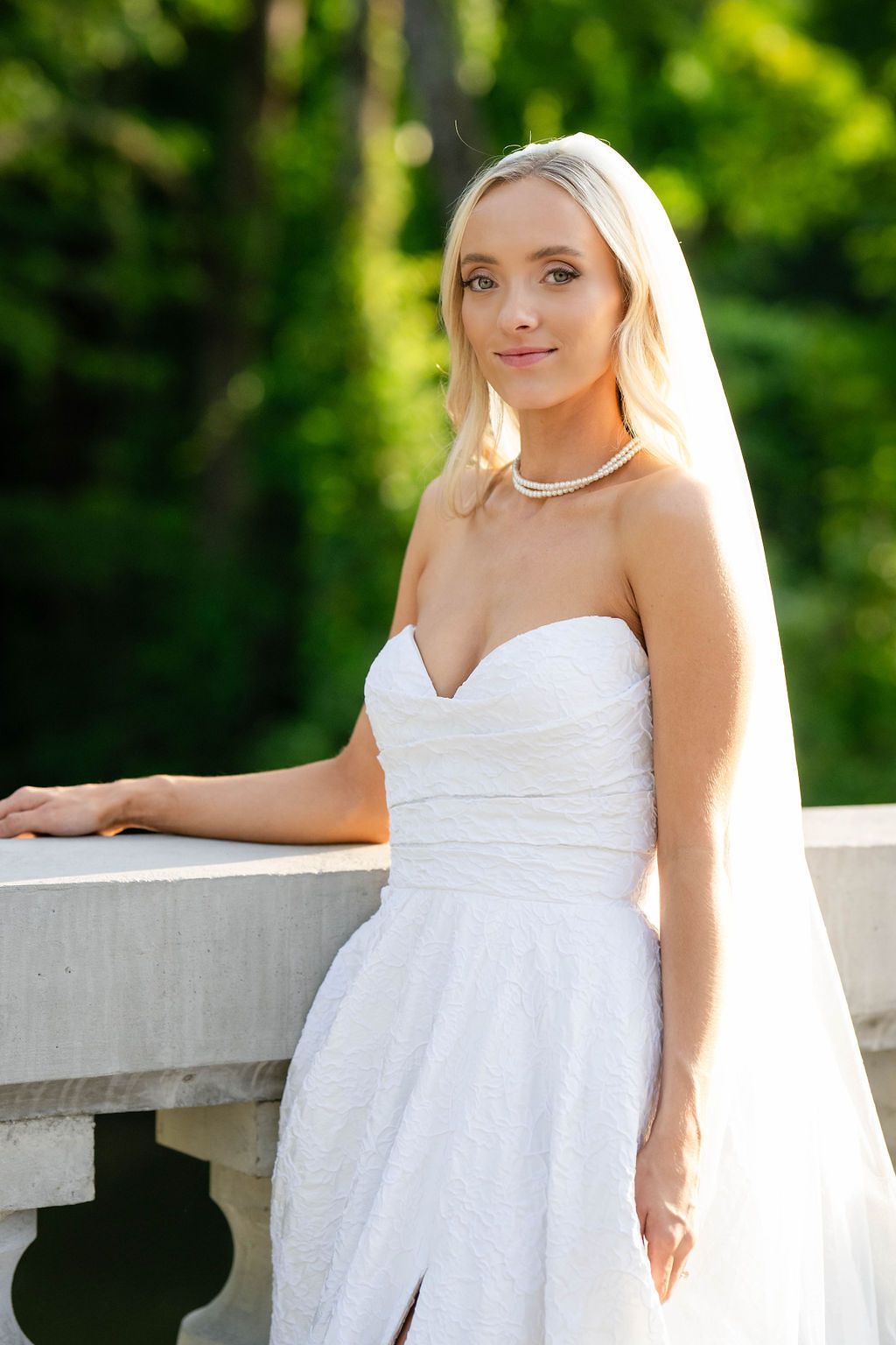 Elegant blonde bride in strapless white lace gown and pearl necklace posing by stone balustrade in garden.