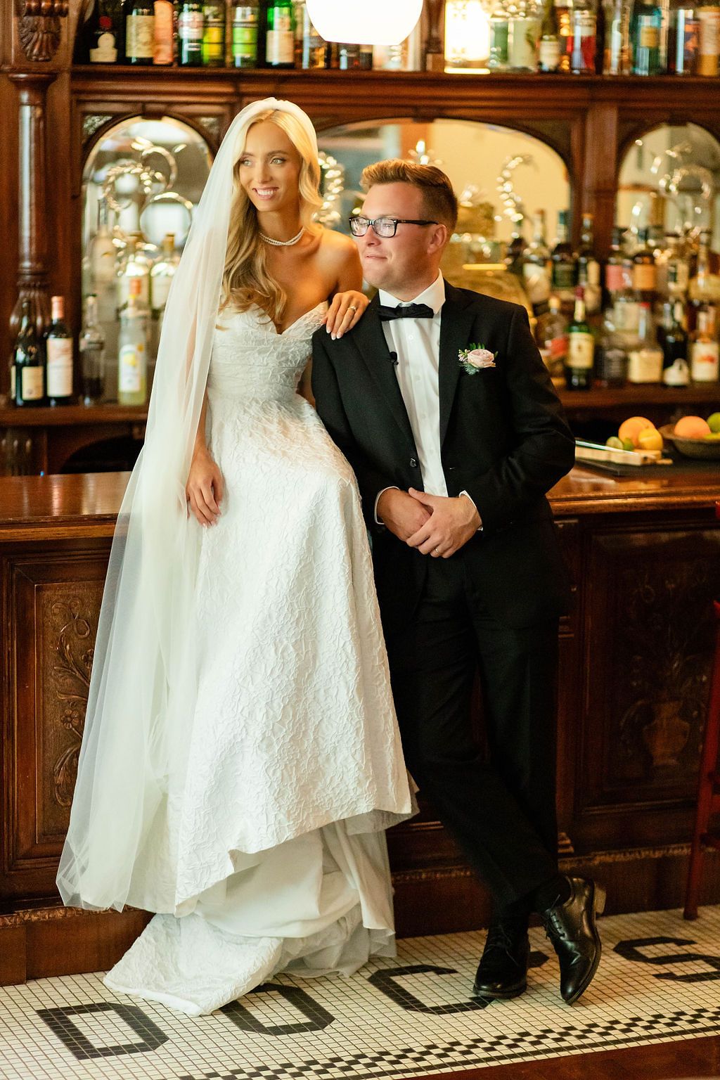 Elegant bride in white lace gown with veil stands beside groom in black tuxedo at a classic wooden bar.