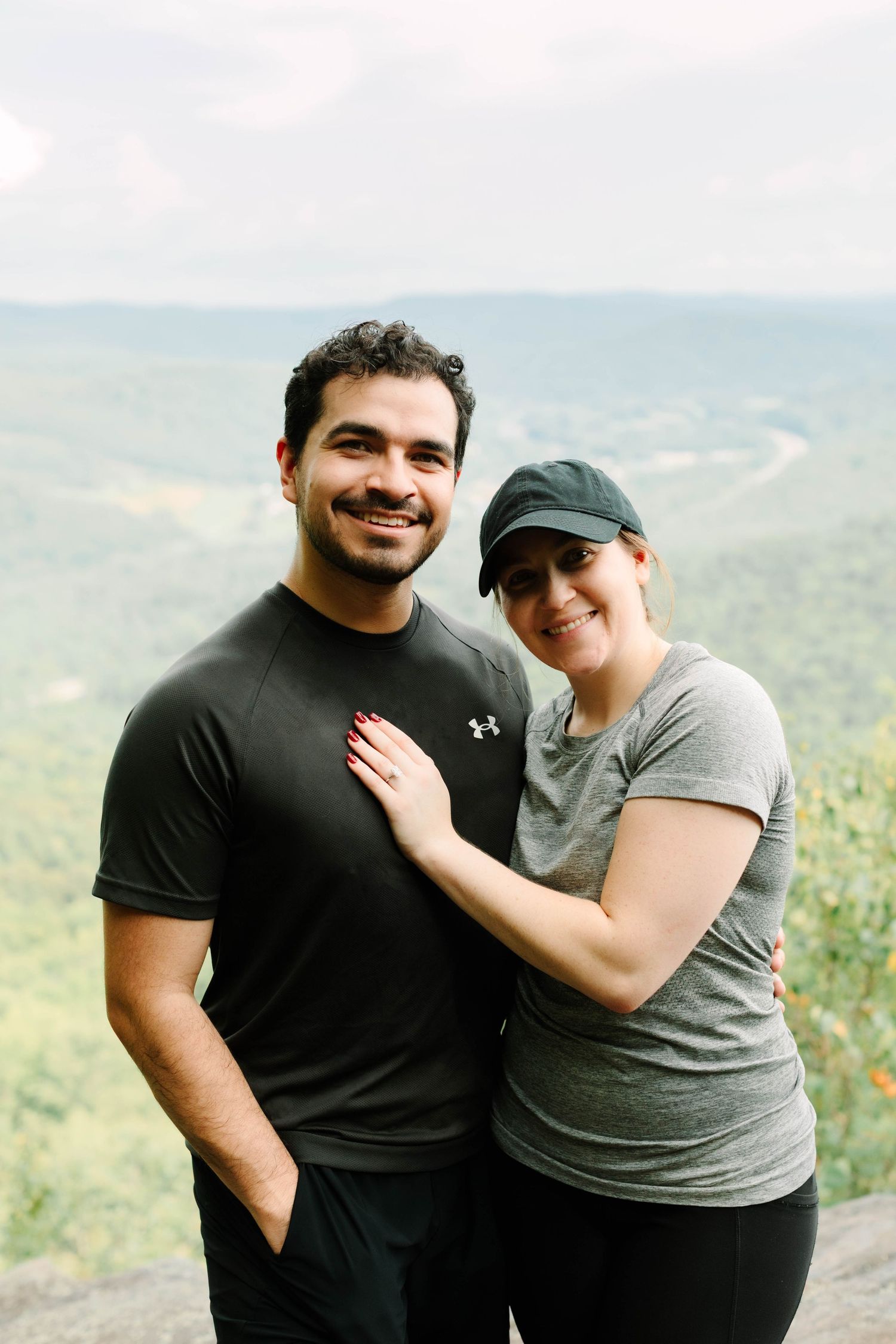 Smiling couple embracing on a rocky overlook with lush green mountain valley views behind them.