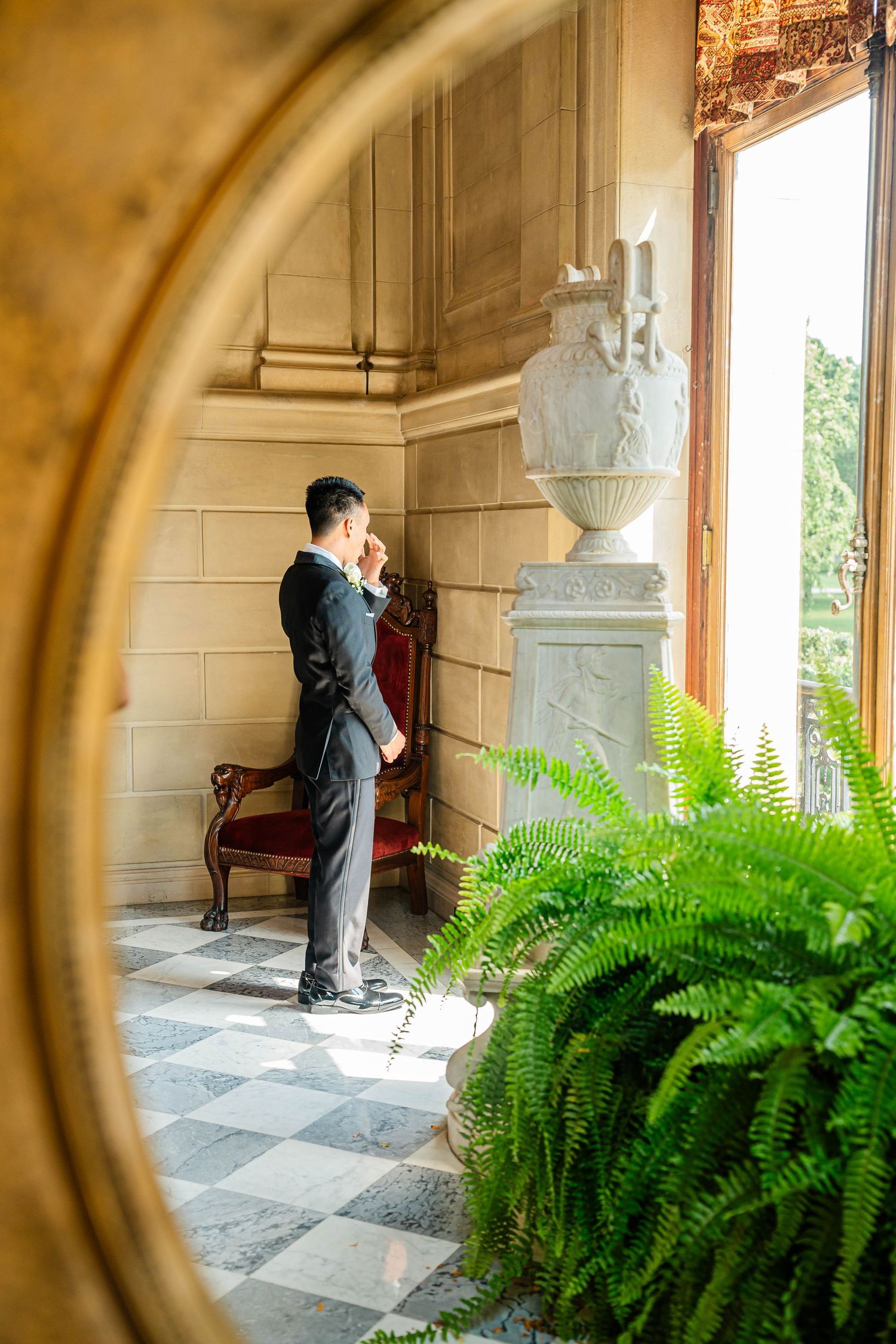 Groom in black tuxedo reflected in ornate mirror inside luxurious marble-floored mansion with ferns.