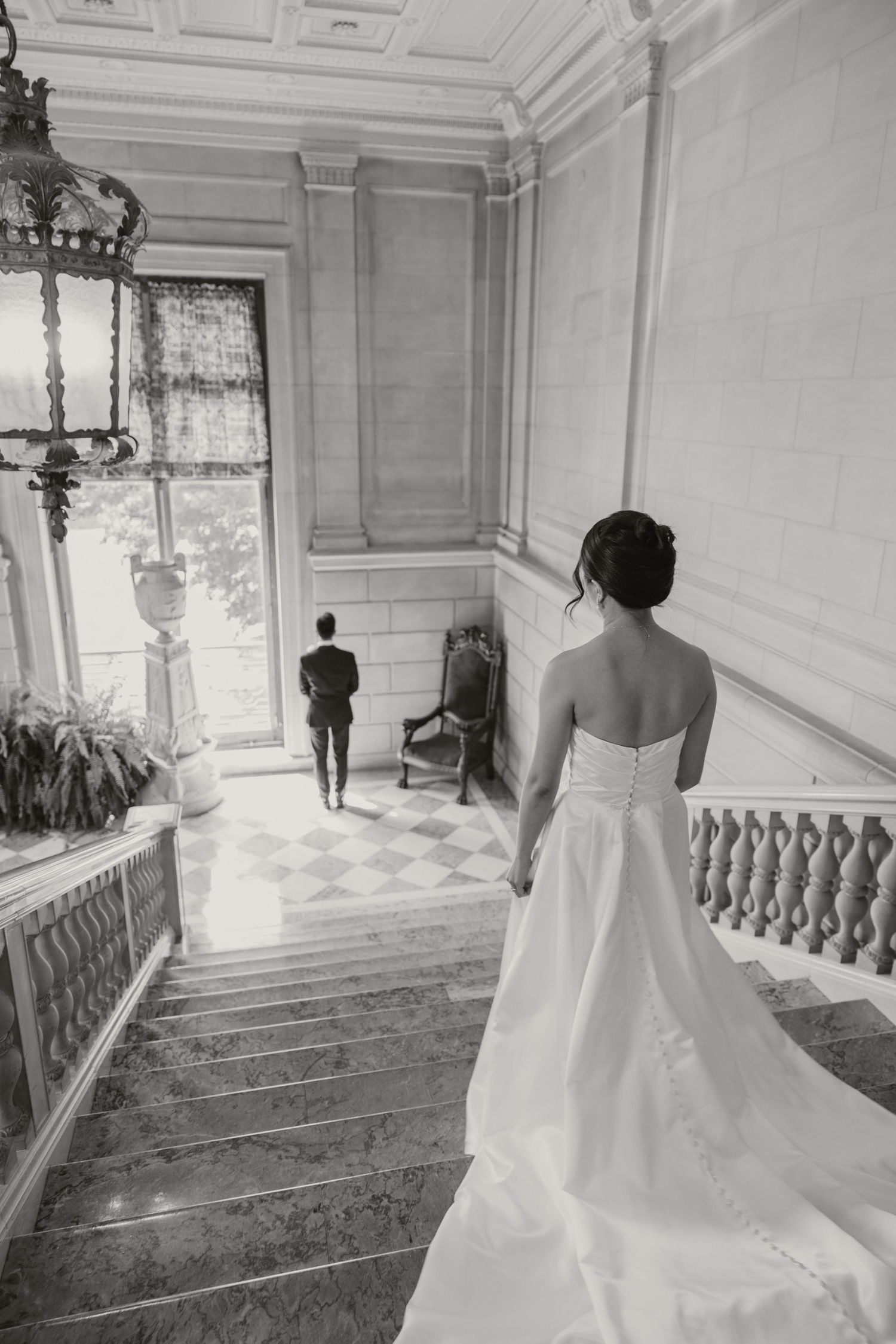 Bride in flowing white gown descending grand staircase toward groom in black and white photo.