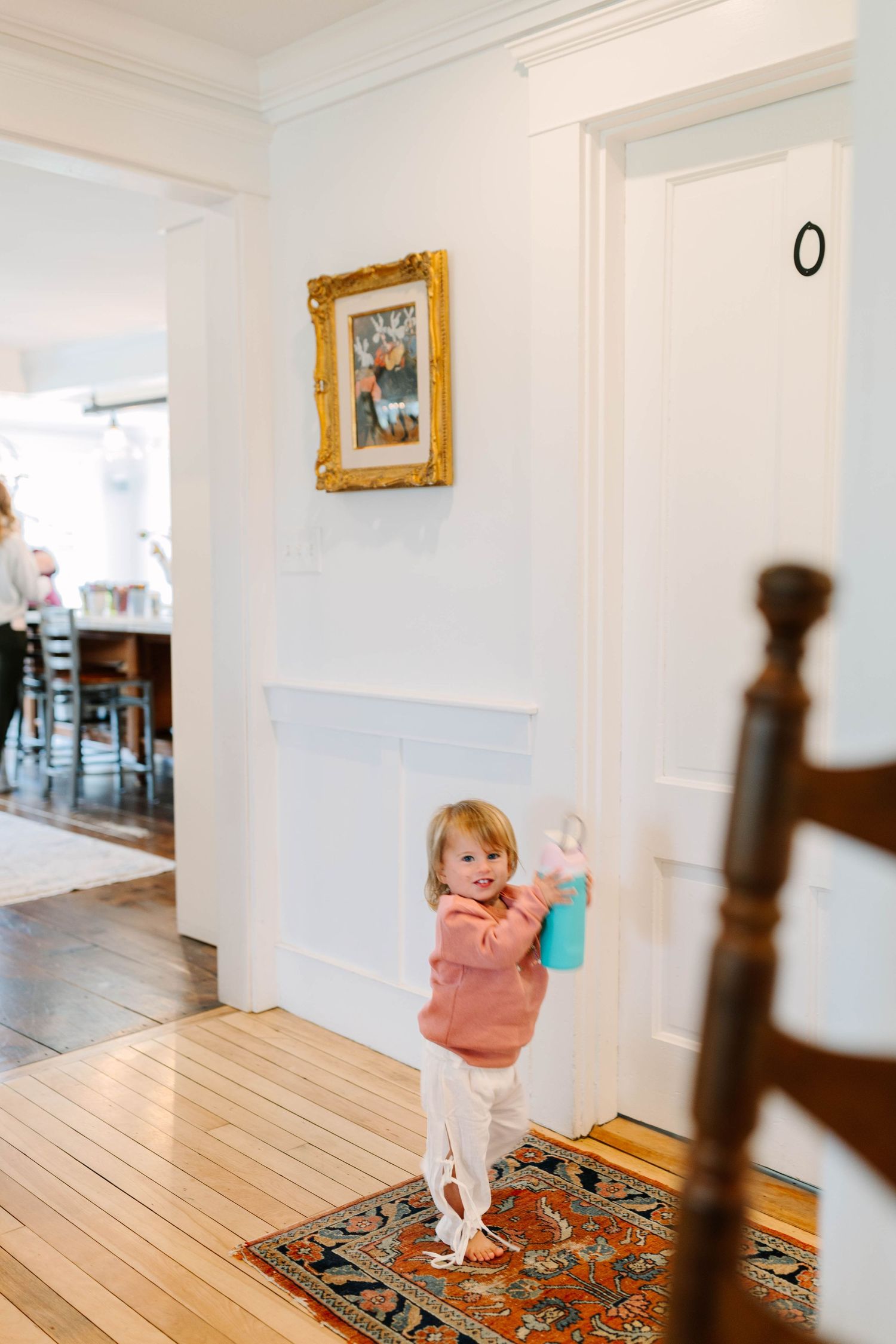 Smiling toddler girl in pink sweater holding a water bottle standing on a colorful rug in a bright white hallway.