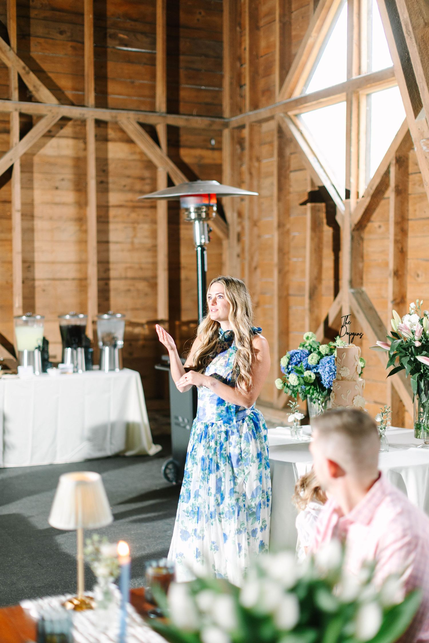 Woman in floral dress speaking at rustic barn wedding reception with blue hydrangea floral arrangements.