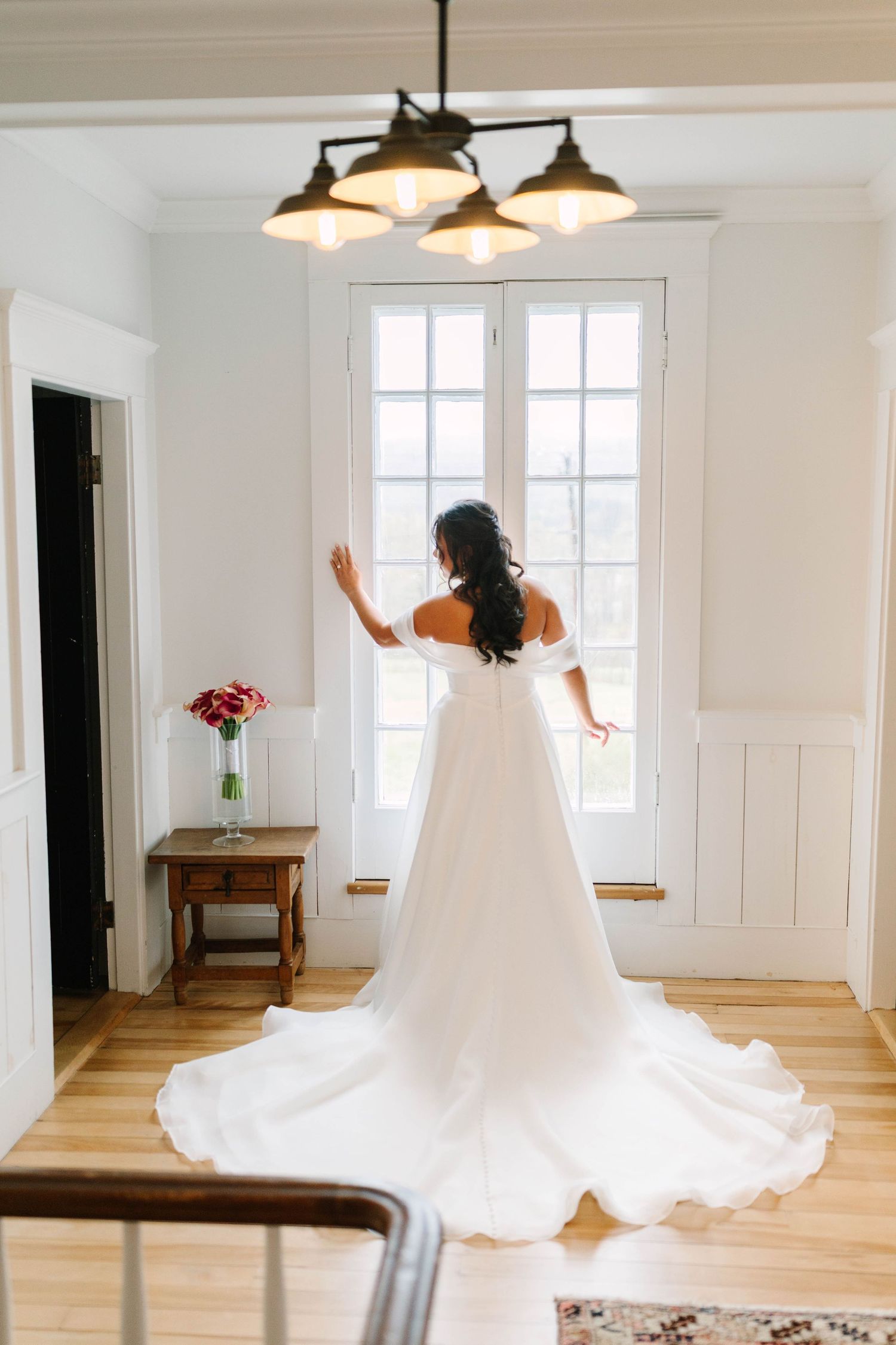 Bride in off-shoulder white gown with long train stands before French doors in bright white room.