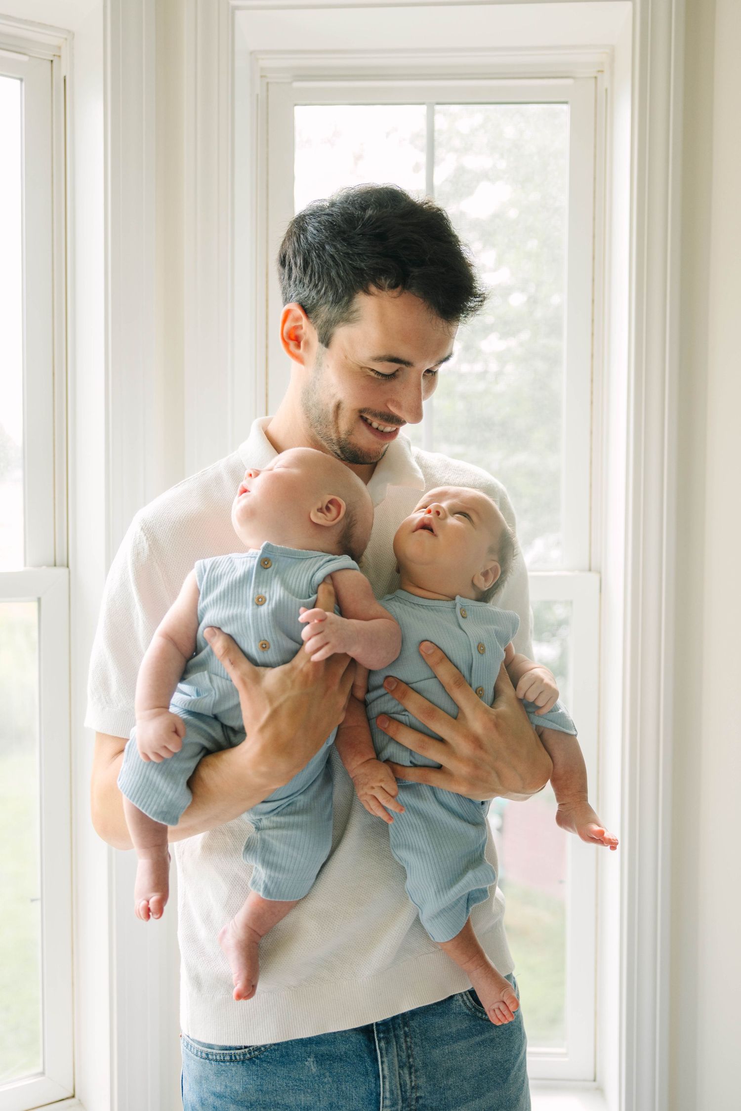 Smiling father holding twin newborn babies in matching blue rompers near bright window.