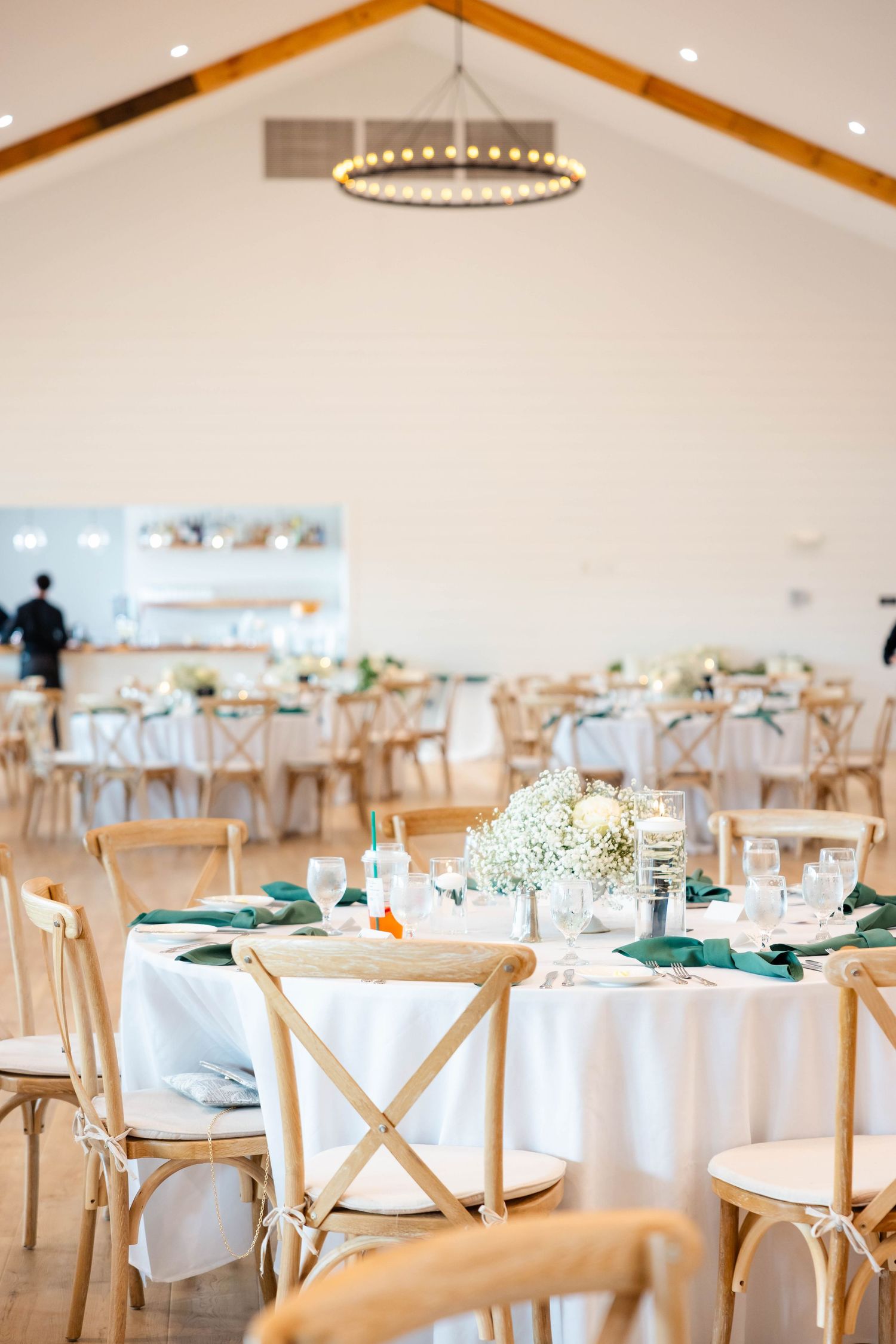Bright wedding reception hall with wooden cross-back chairs, green napkins, and baby's breath centerpieces.