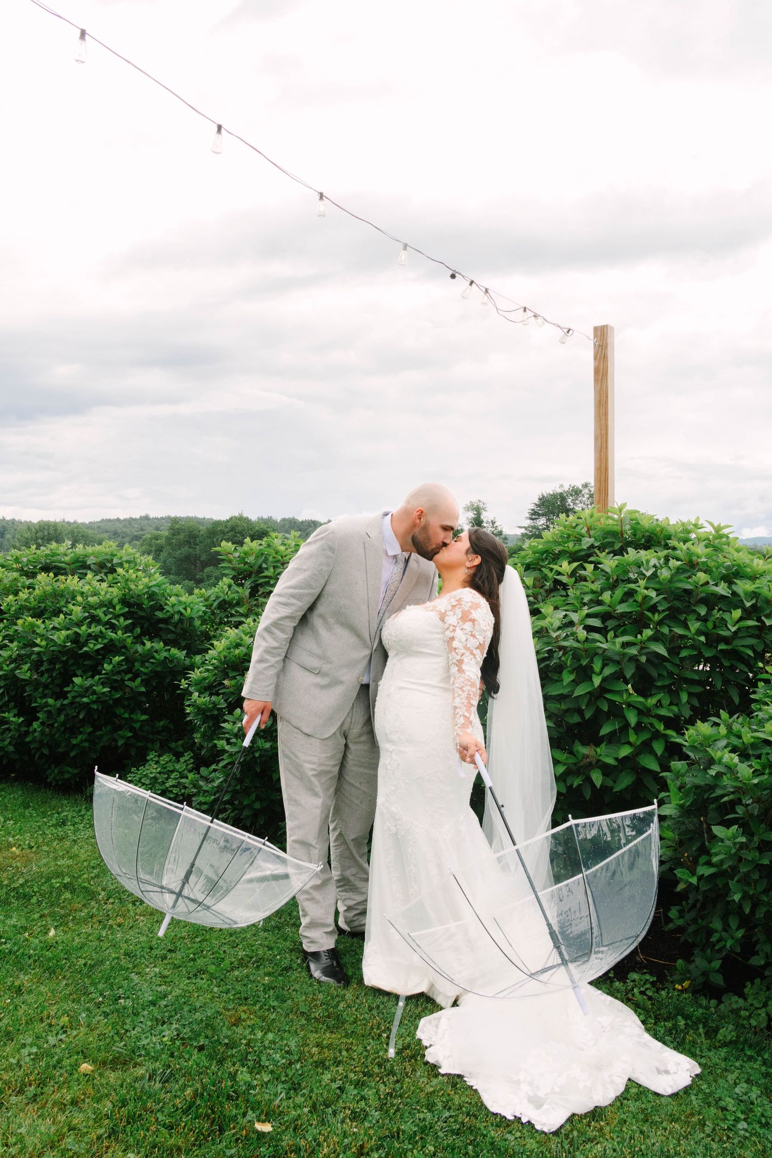 Wedding couple shares a kiss outdoors with a clear umbrella on the ground and string lights overhead amid green foliage.