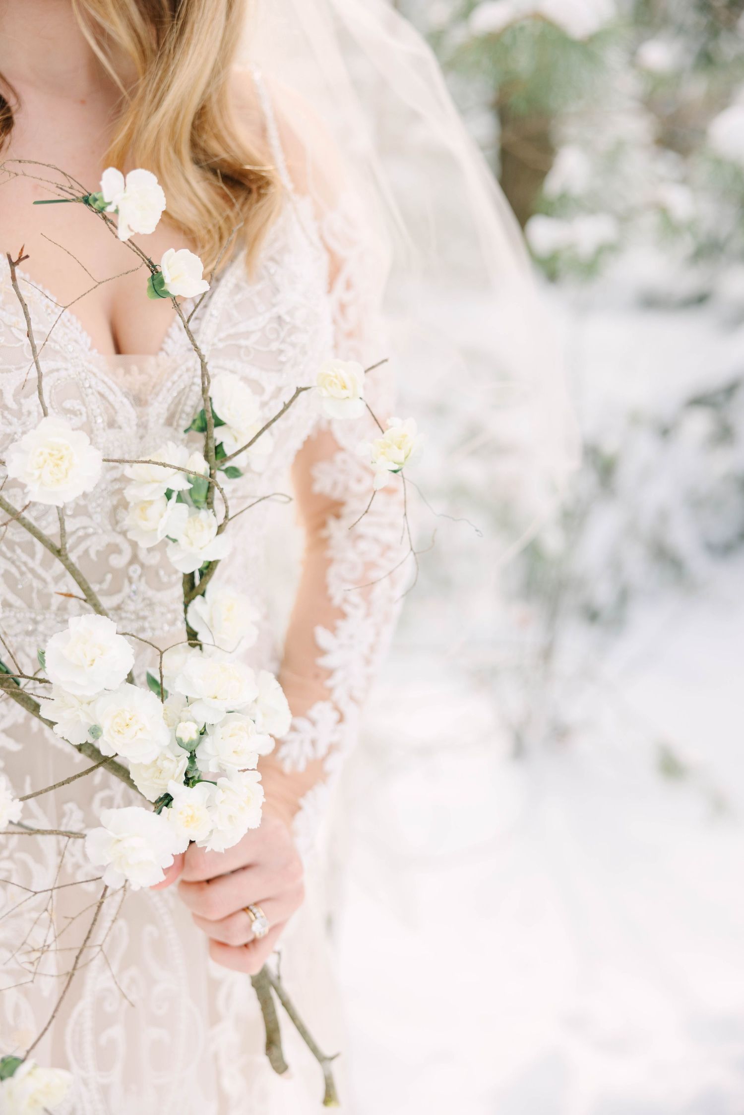 Close-up of a bride in lace gown holding white floral branches with small roses in a snowy winter setting.