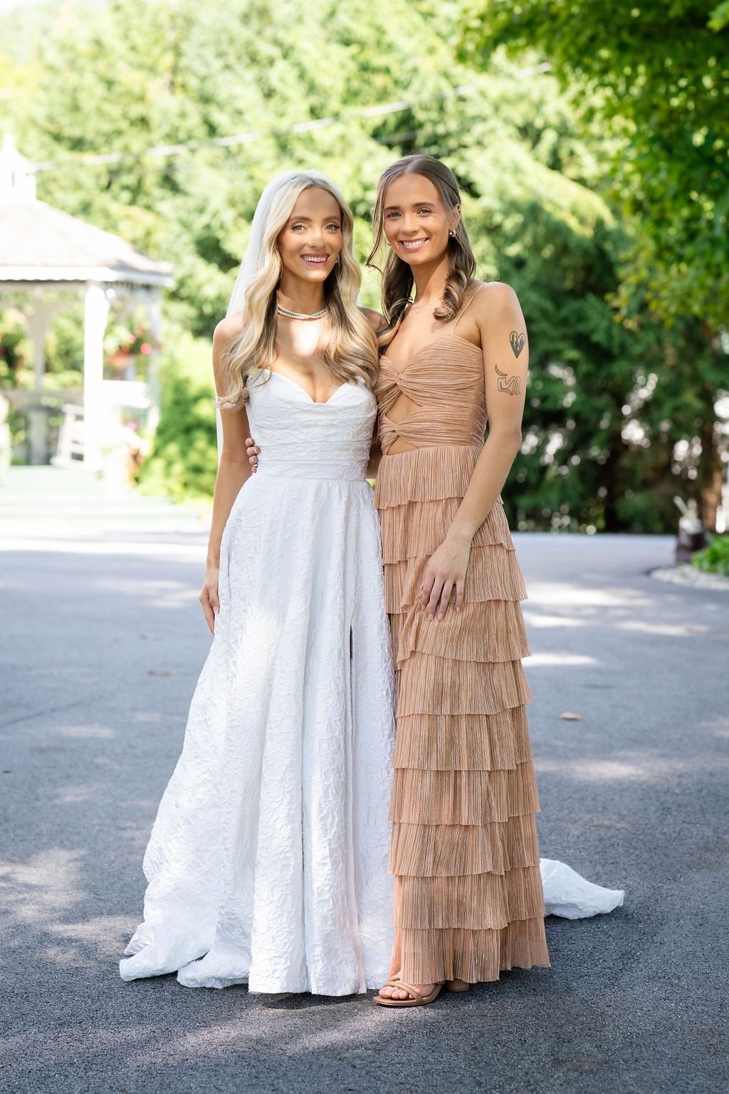 Bride in white lace gown poses with a bridesmaid in a tan ruffled dress in a sunlit outdoor setting.