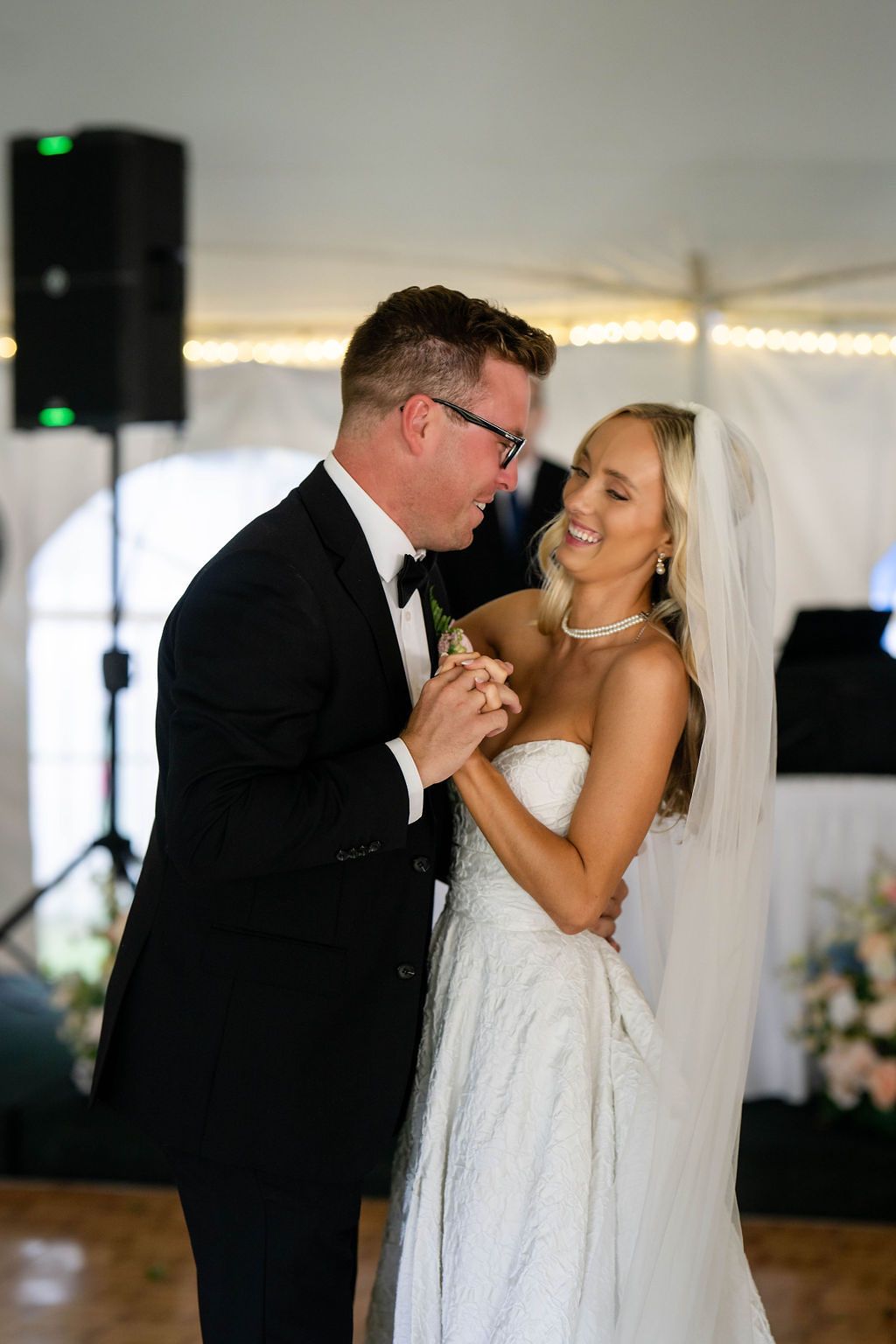 Bride and groom share their first dance, smiling joyfully under warm lights in a white wedding tent.