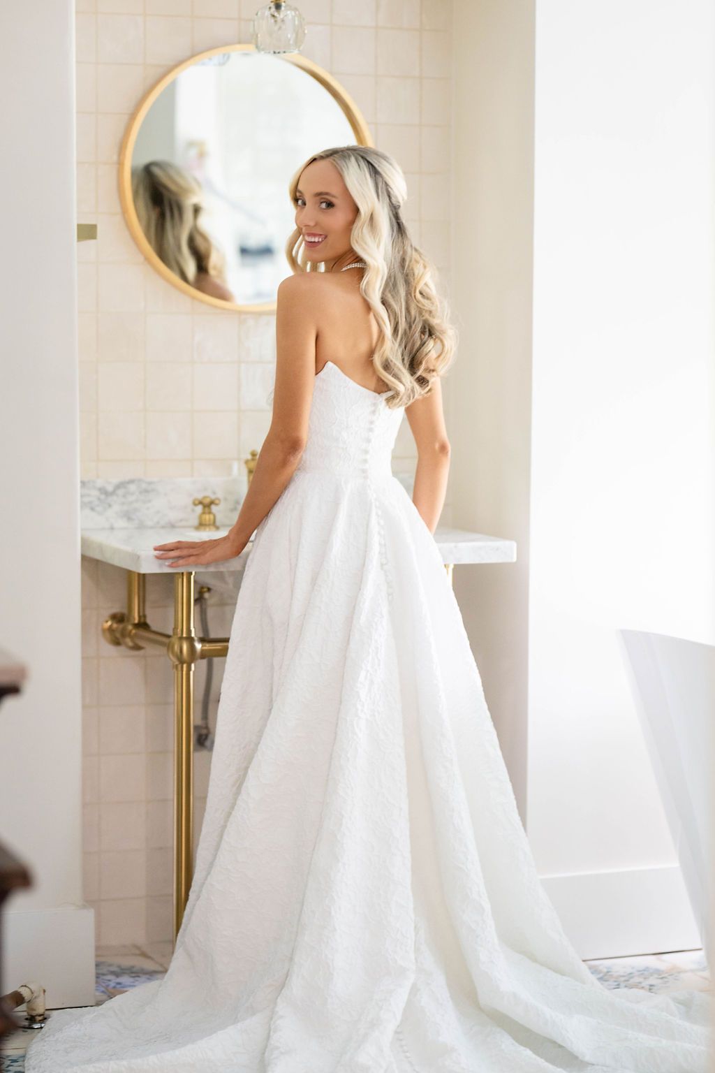 Smiling bride in strapless white ball gown with lace detail poses in elegant bathroom with gold fixtures and round mirror.