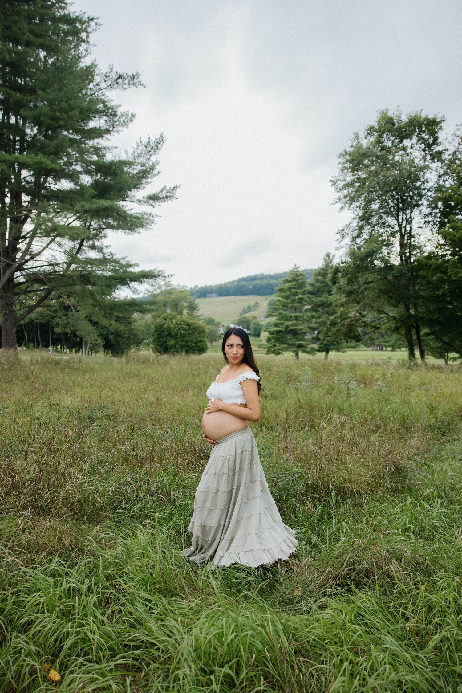 Pregnant woman in white crop top and gray maxi skirt standing in lush green meadow surrounded by trees.