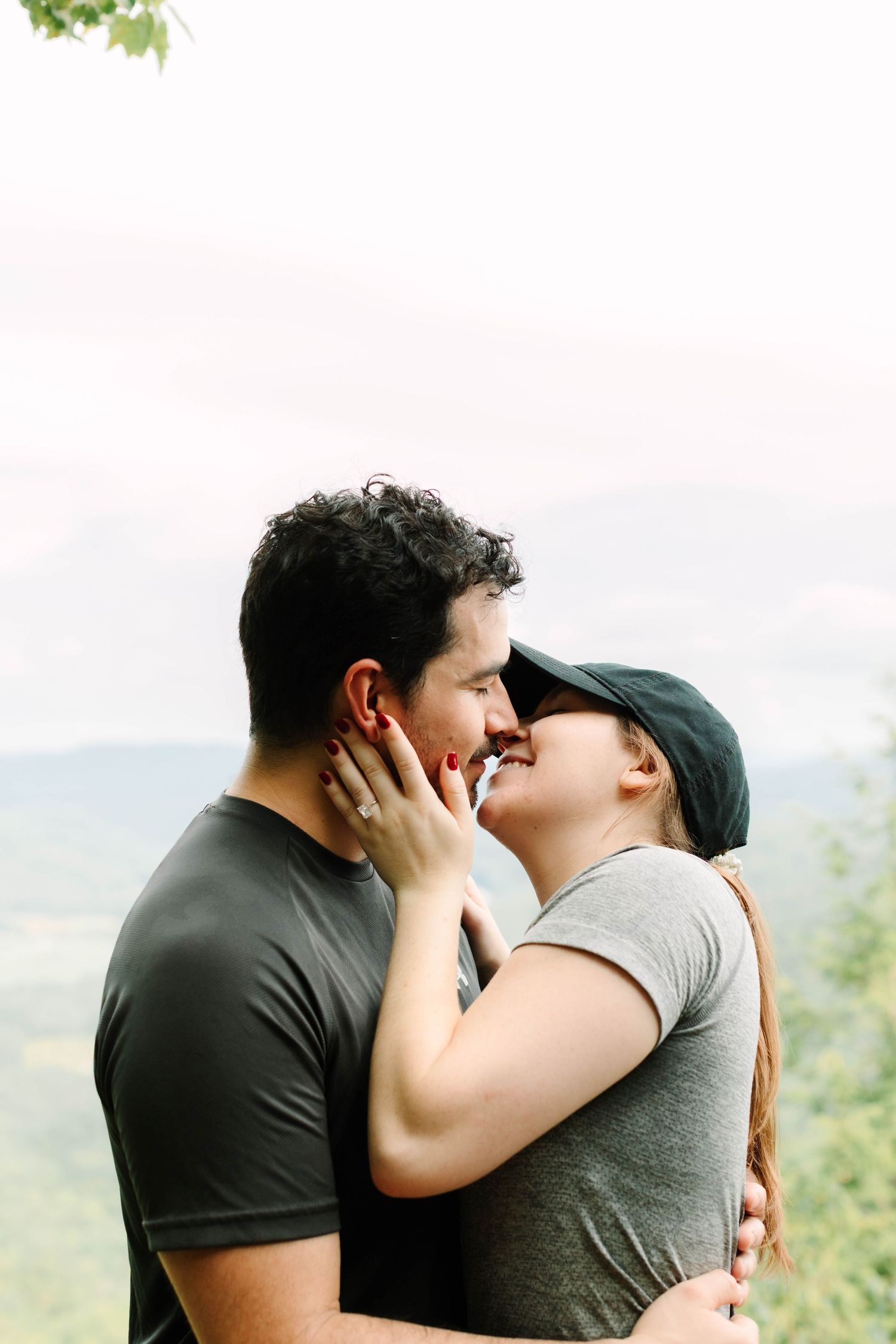 Couple sharing a romantic almost-kiss outdoors, woman wearing cap and engagement ring, mountains visible in background.