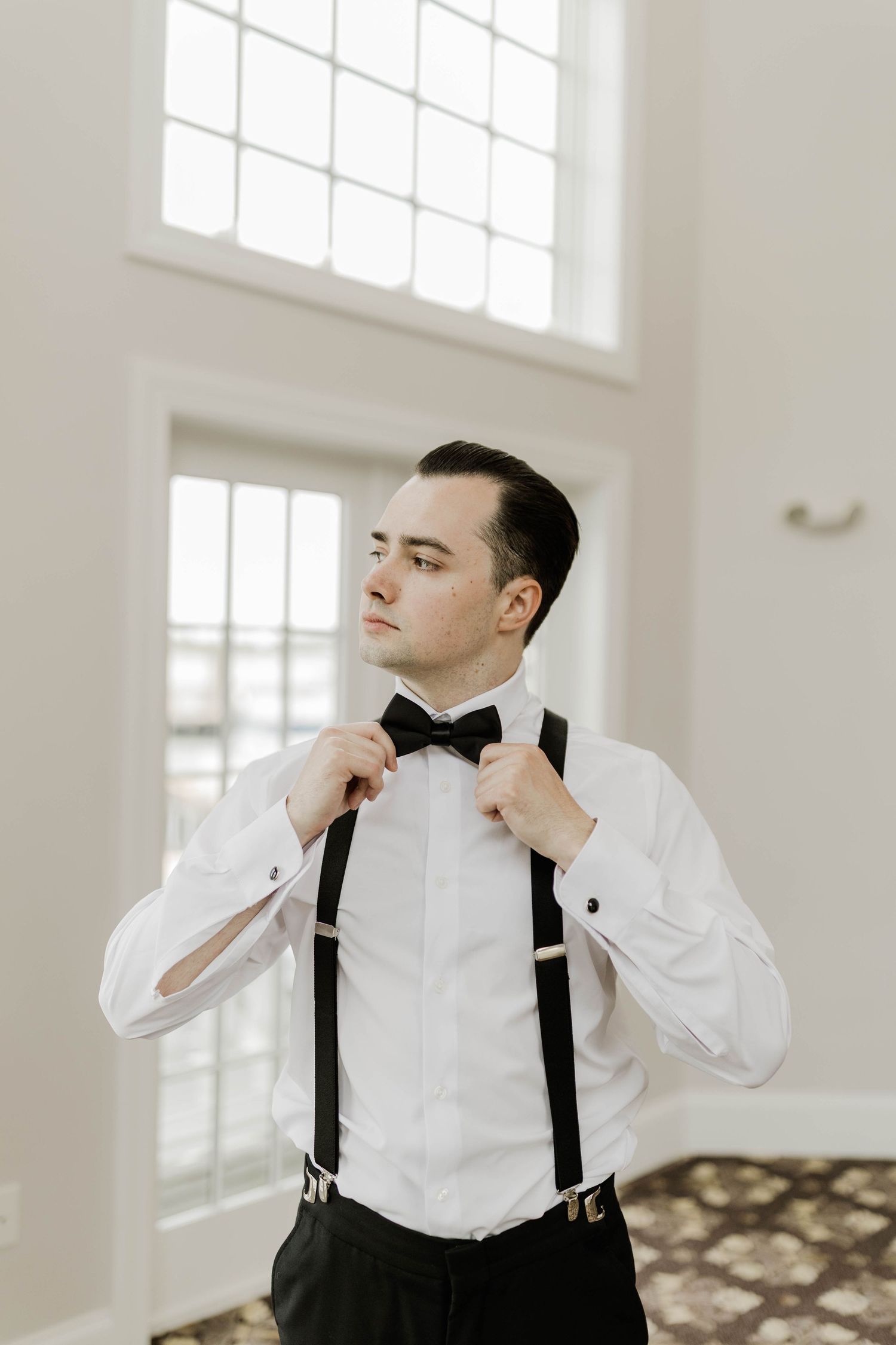 Dapper groom in white shirt adjusting black bow tie with suspenders before wedding ceremony.