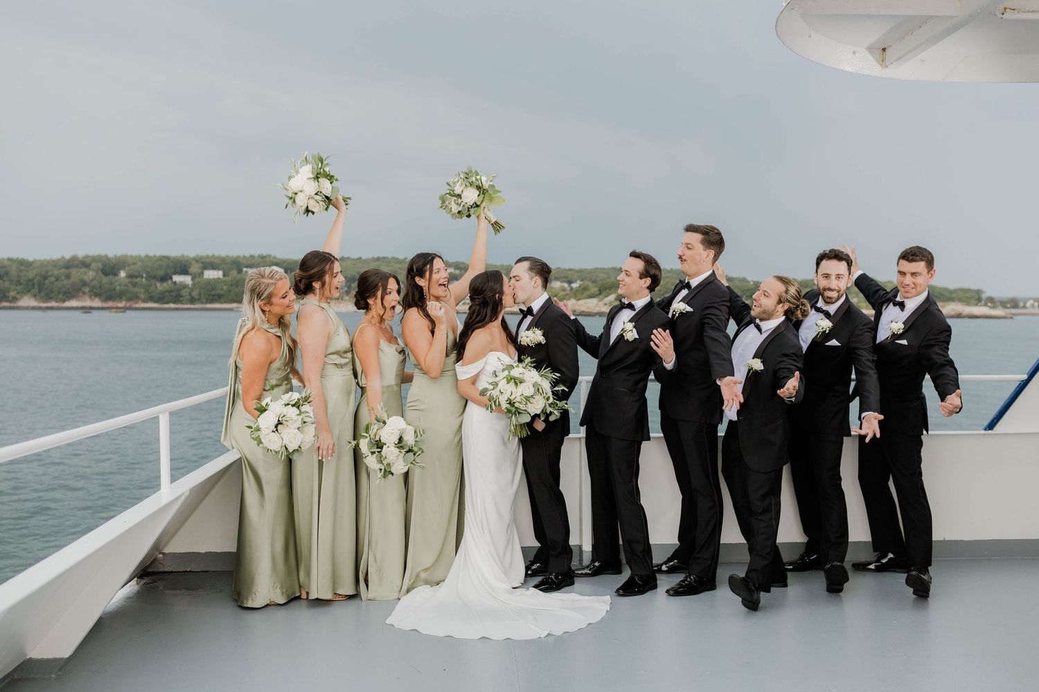 Wedding party poses on yacht deck with bridesmaids in sage green and groomsmen in black tuxedos.