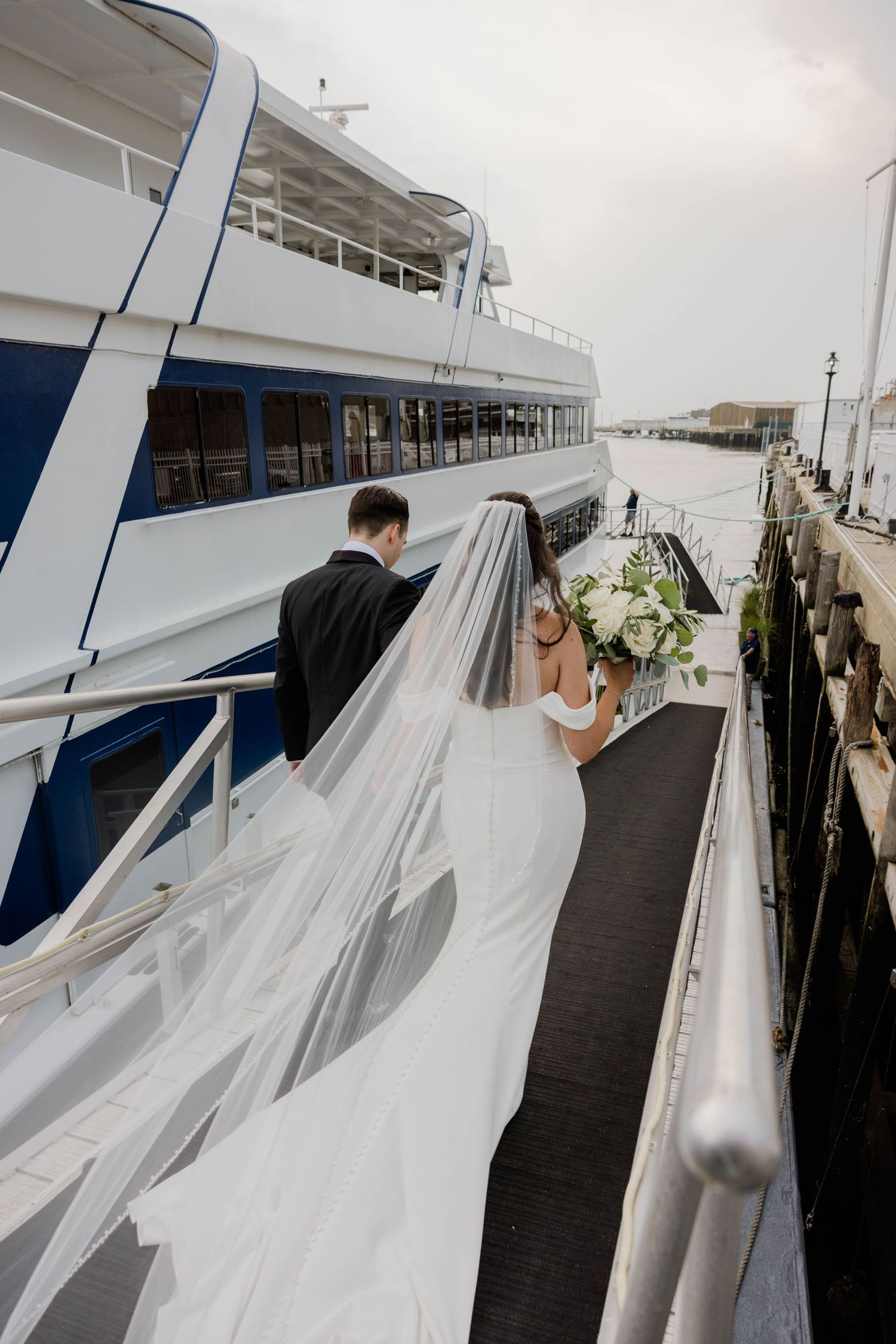 Bride and groom in wedding attire boarding a luxury yacht at a marina.