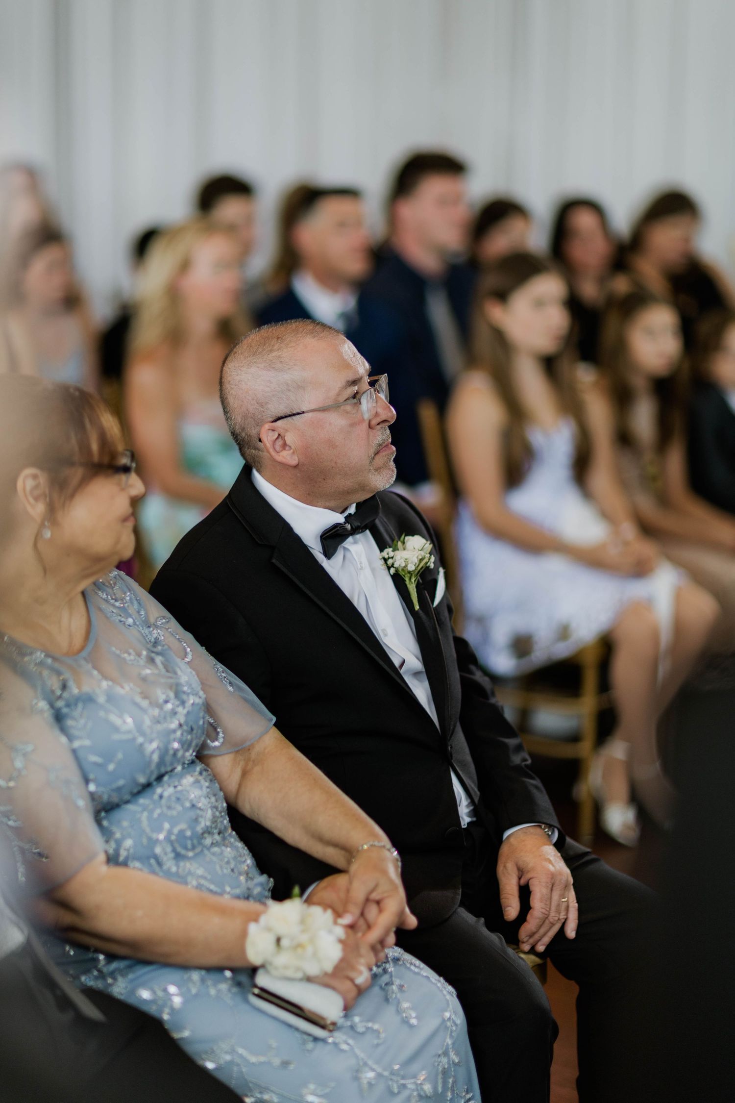 Wedding guests in formal attire seated during a ceremony, father of groom watching.