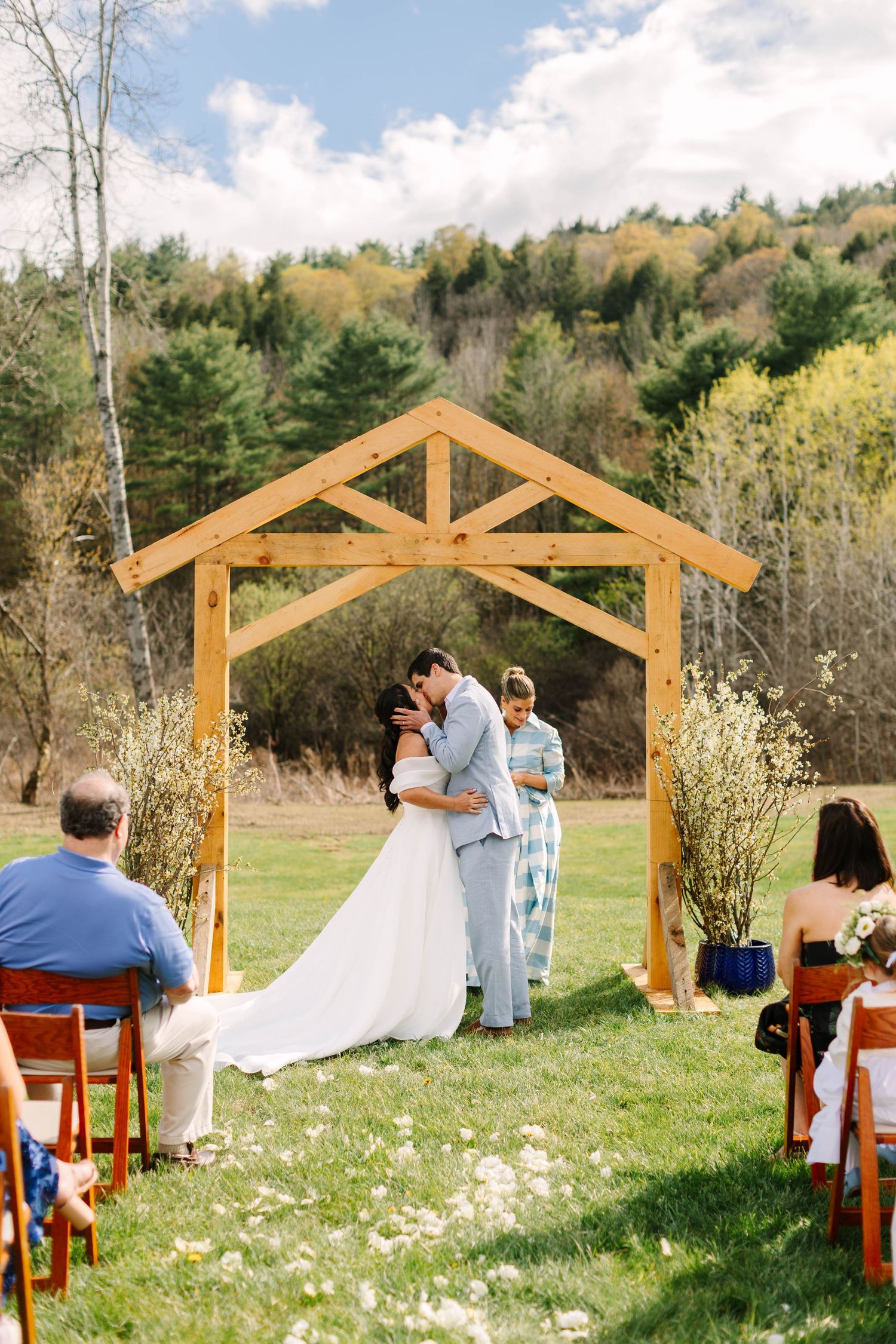 Couple shares first kiss under wooden arch during outdoor wedding ceremony with autumn hills backdrop.