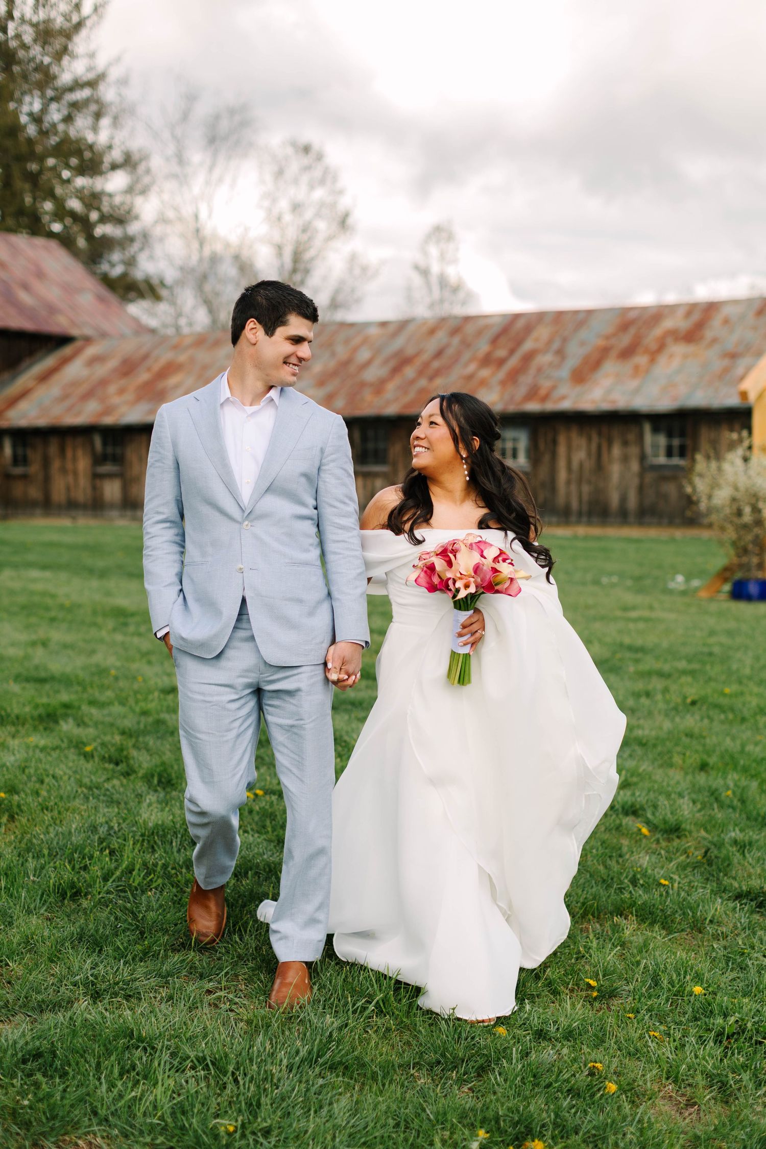 Bride and groom walking hand in hand on green grass with rustic barn backdrop on their wedding day.