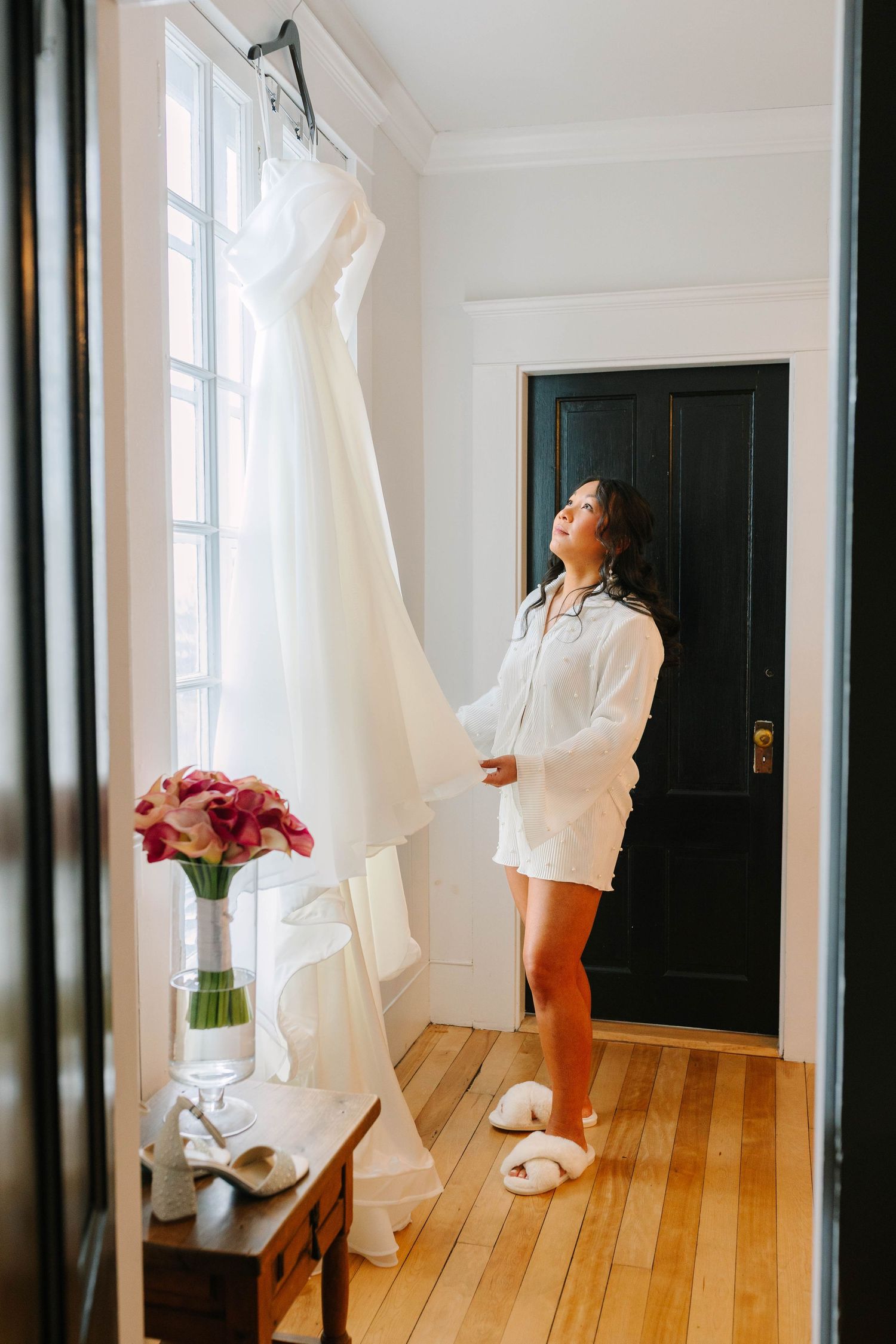 Bride in white shirt admiring her wedding dress hanging by a window with bridal shoes and flowers nearby.