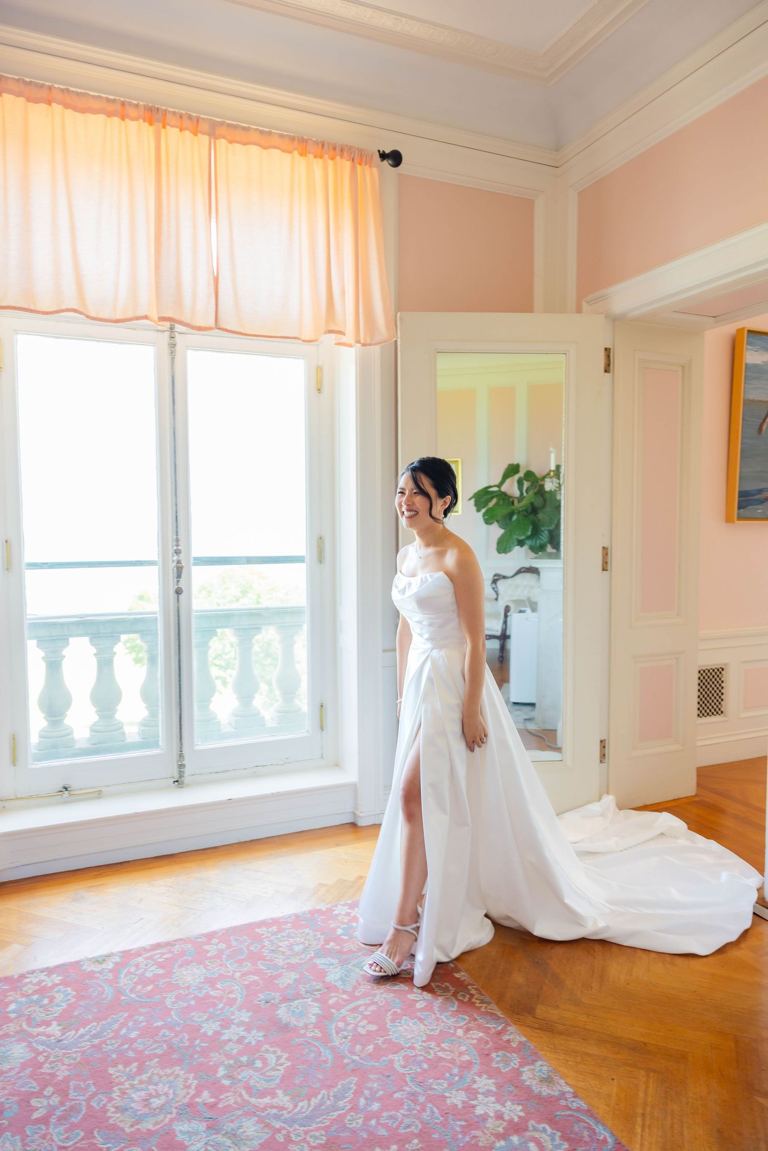 Elegant bride in a white halter gown with a long train stands by a sunlit window in a pink-walled room.