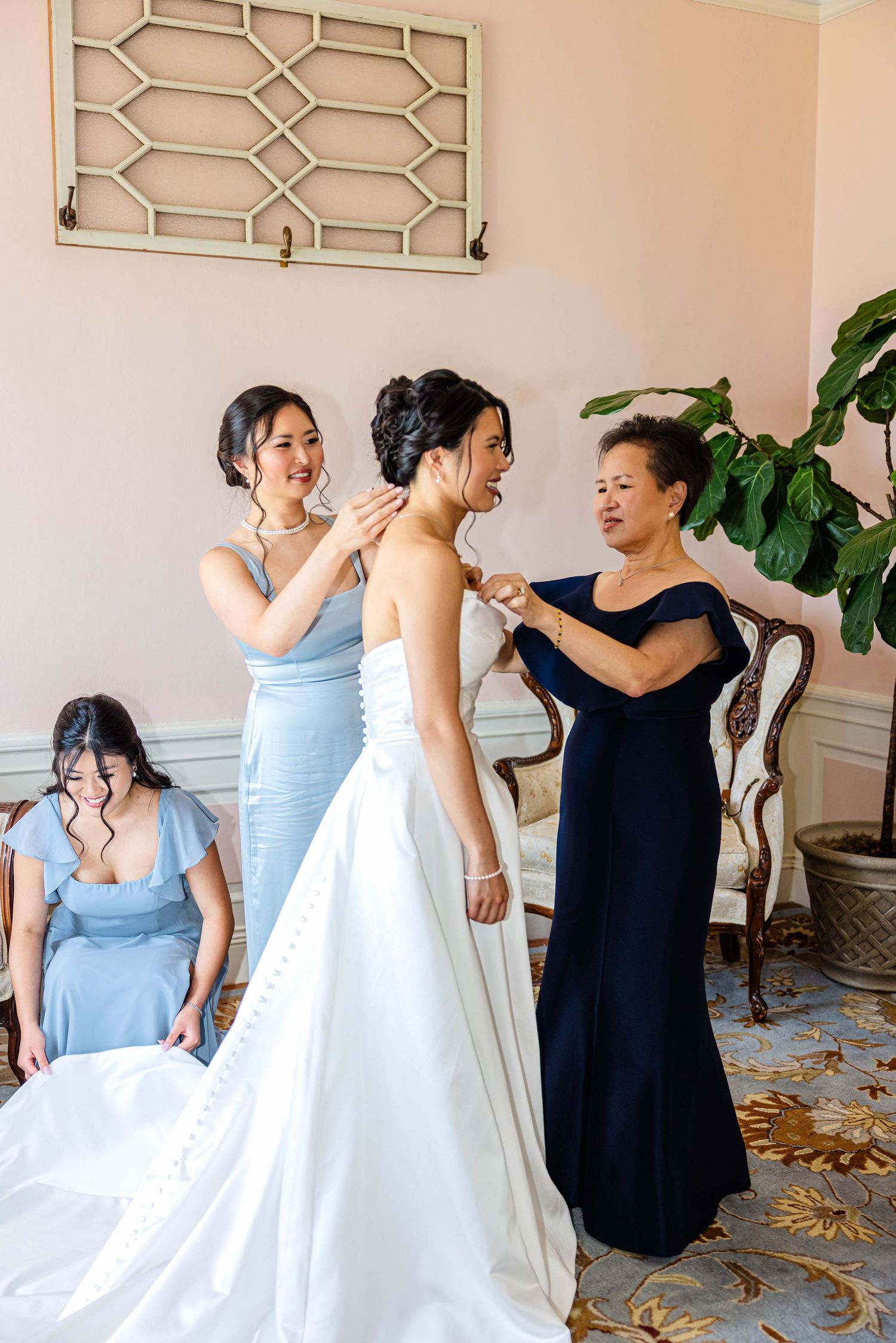 Bridesmaids in blue dresses help bride into white wedding gown in elegant pink room.